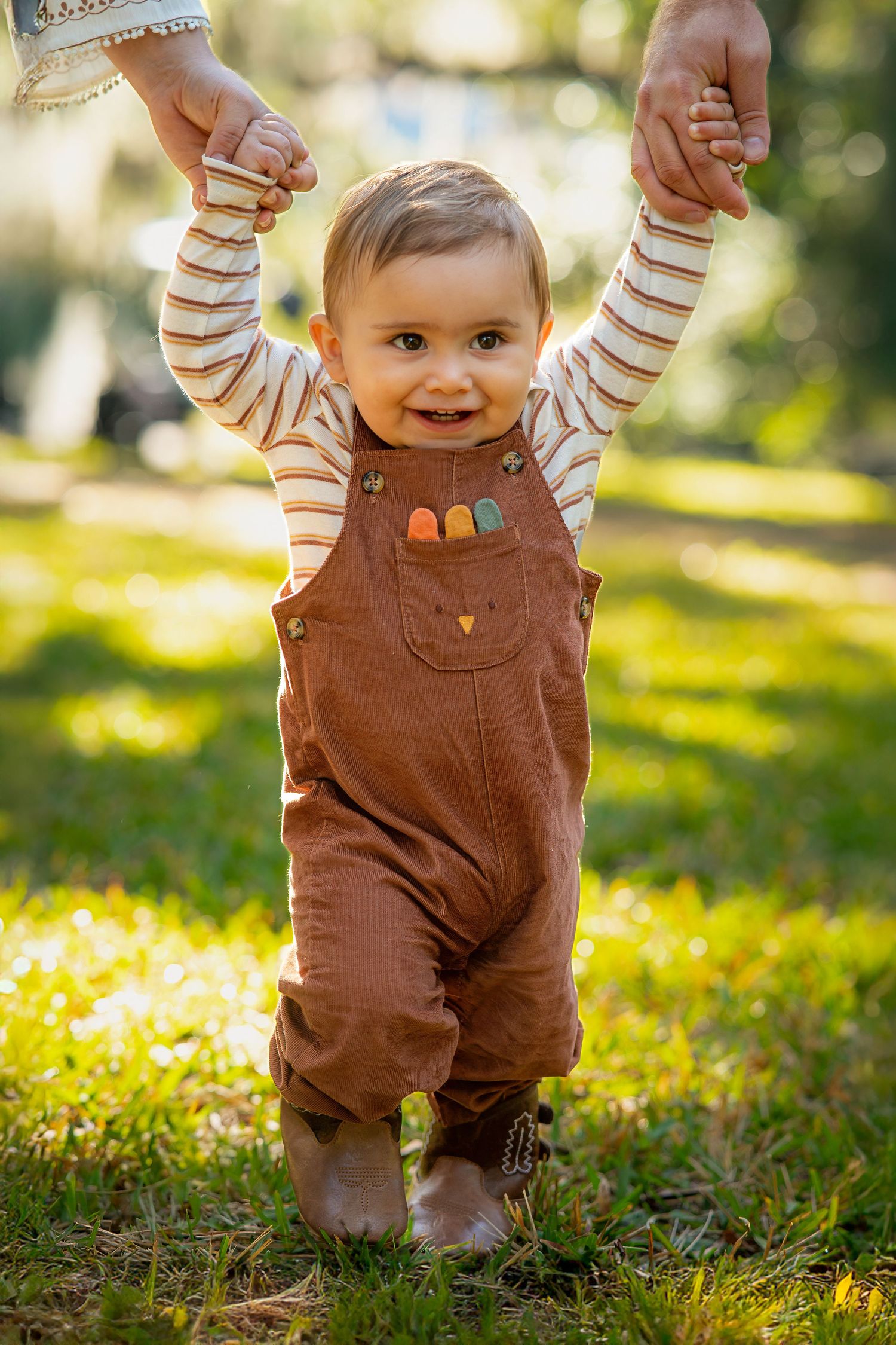 Small child in brown overalls and striped shirt walking on grass in golden sunlight.