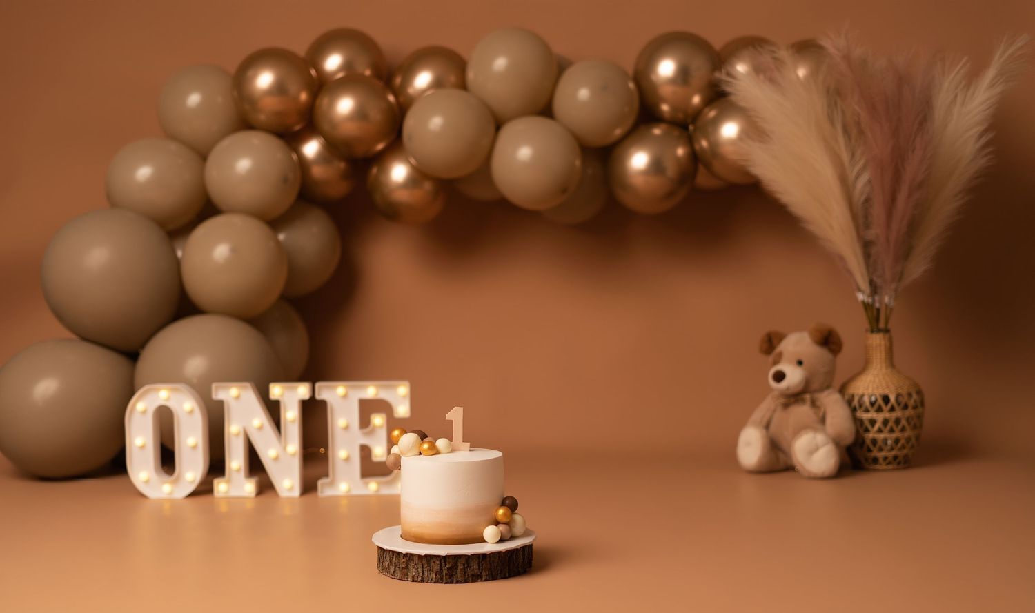 First birthday cake smash setup with ONE marquee letters, teddy bear, pampas grass, and brown balloon garland on beige backdrop.