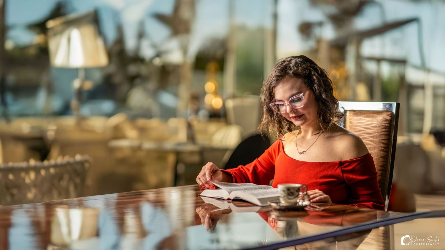 A woman in red reading a book while sitting at an outdoor cafe table in warm sunlight.
