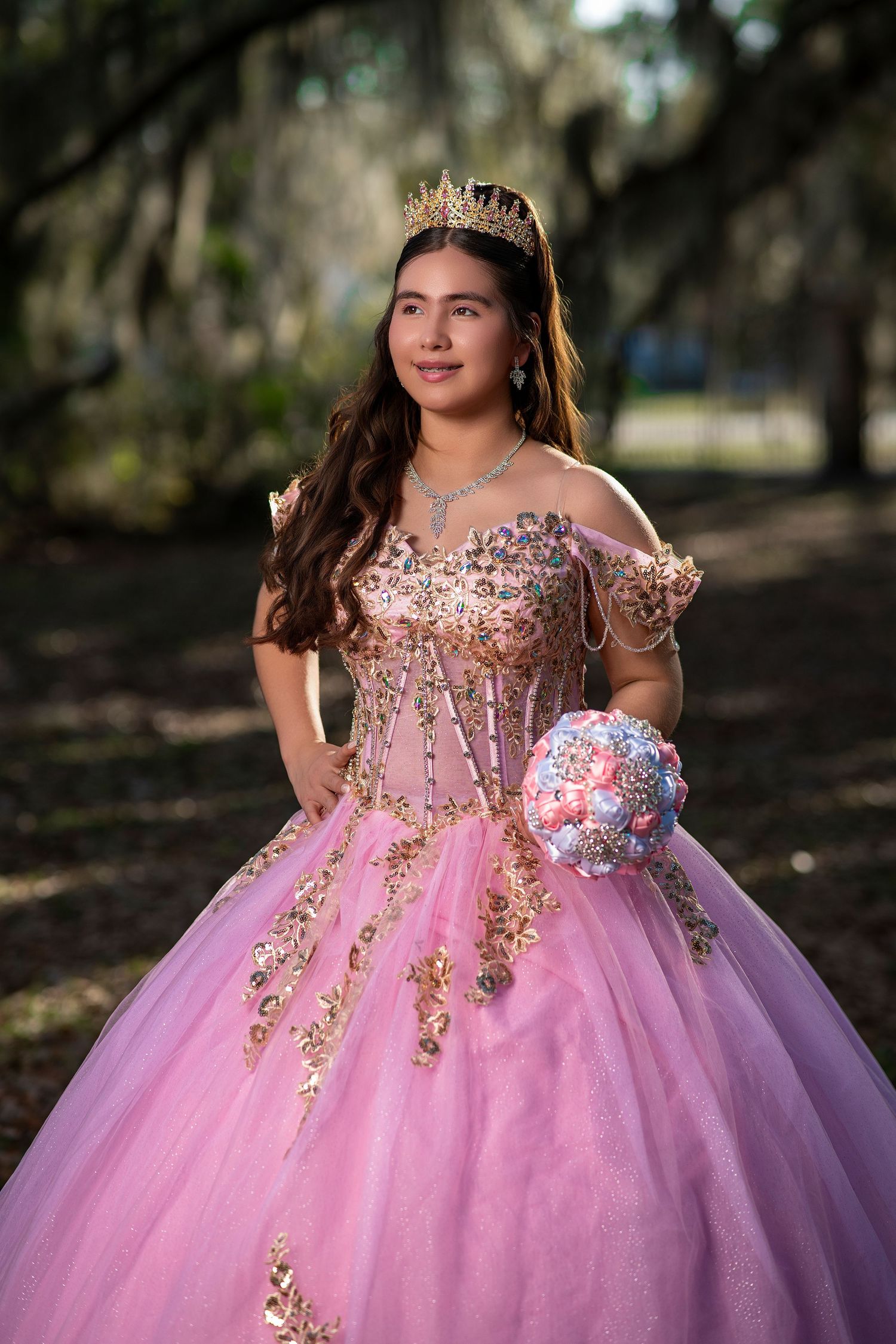 Young woman in pink quinceañera ball gown with gold embroidery, tiara, and jeweled bouquet posing outdoors.