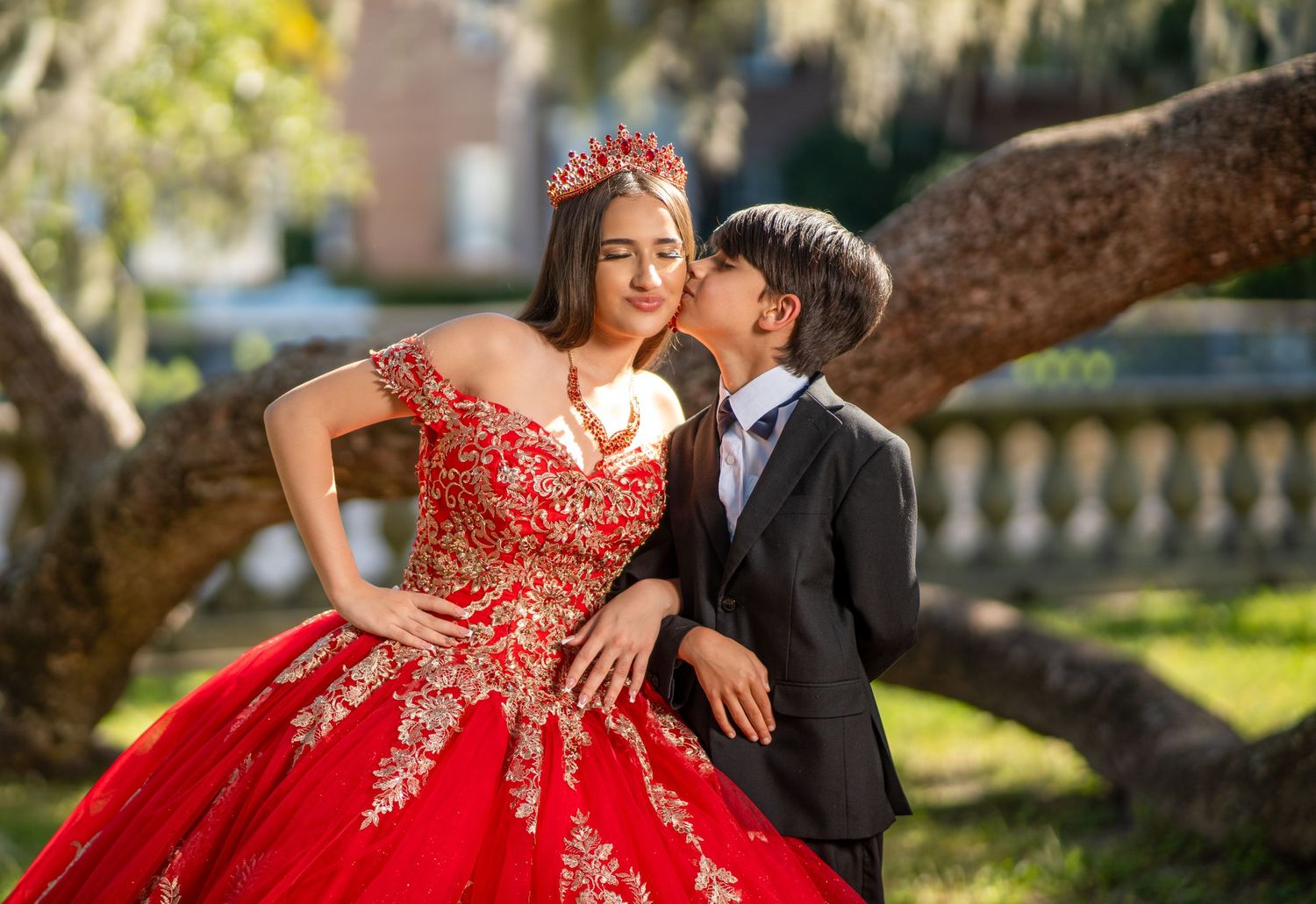 A couple poses for romantic portraits in a garden setting, with the person wearing a red and gold formal gown.