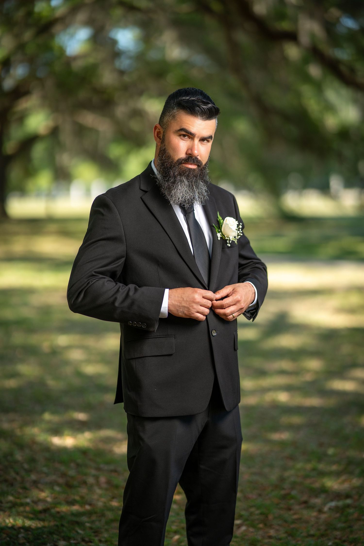Bearded groom in black suit with boutonniere buttoning jacket outdoors in lush green park setting.