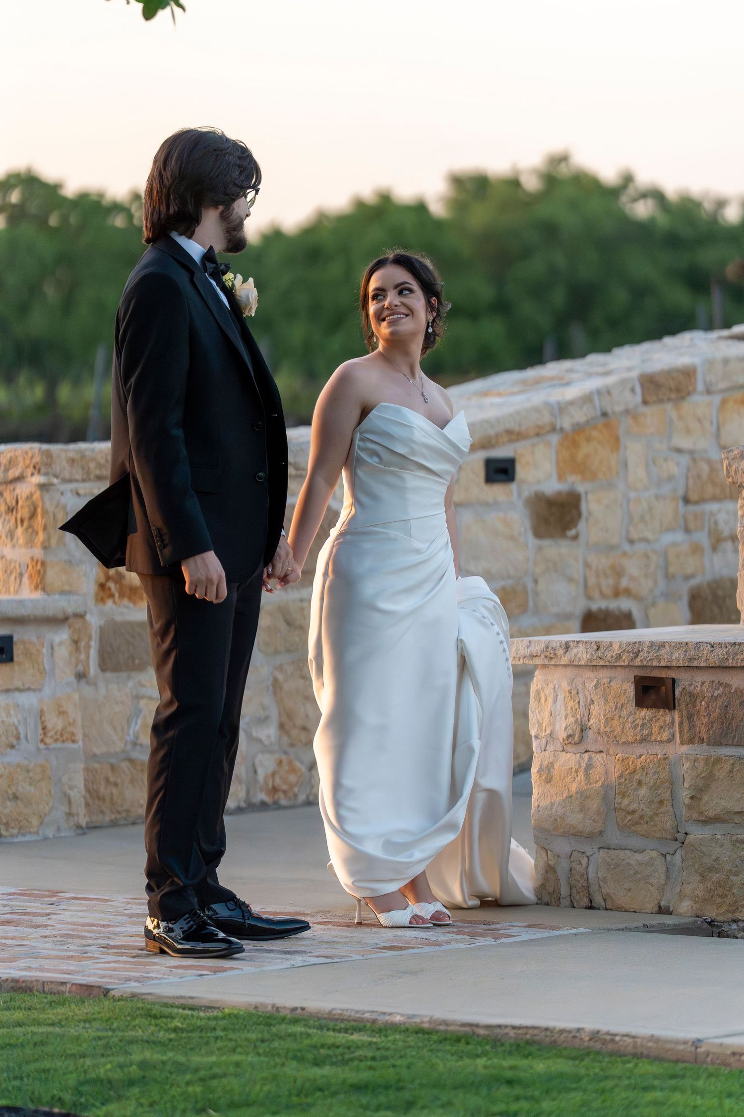 Bride in strapless white gown holds grooms hand outdoors at golden hour near stone wall.