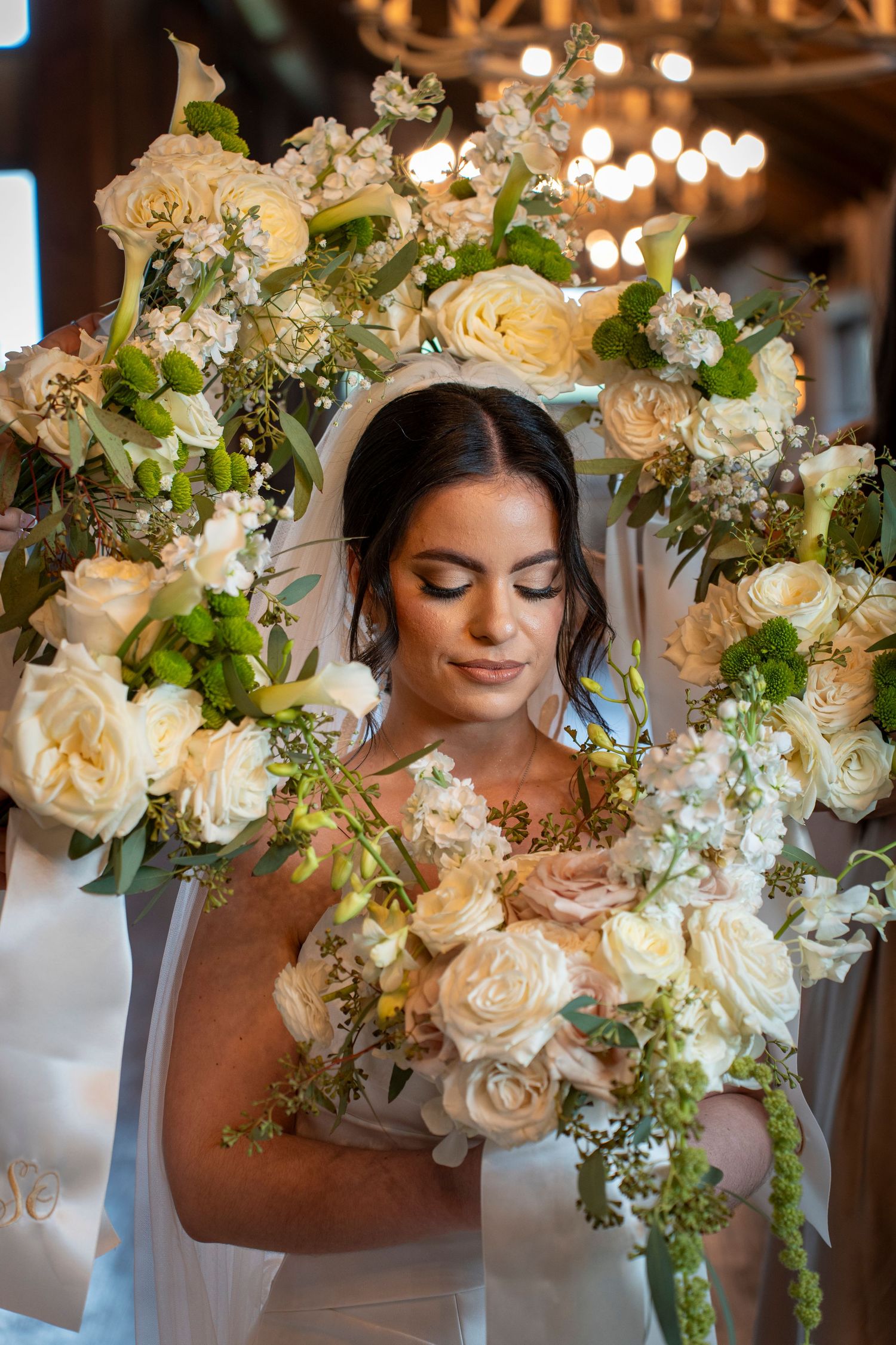 Bride surrounded by lush white and green floral wreath bouquet, eyes closed, wearing veil at elegant wedding.