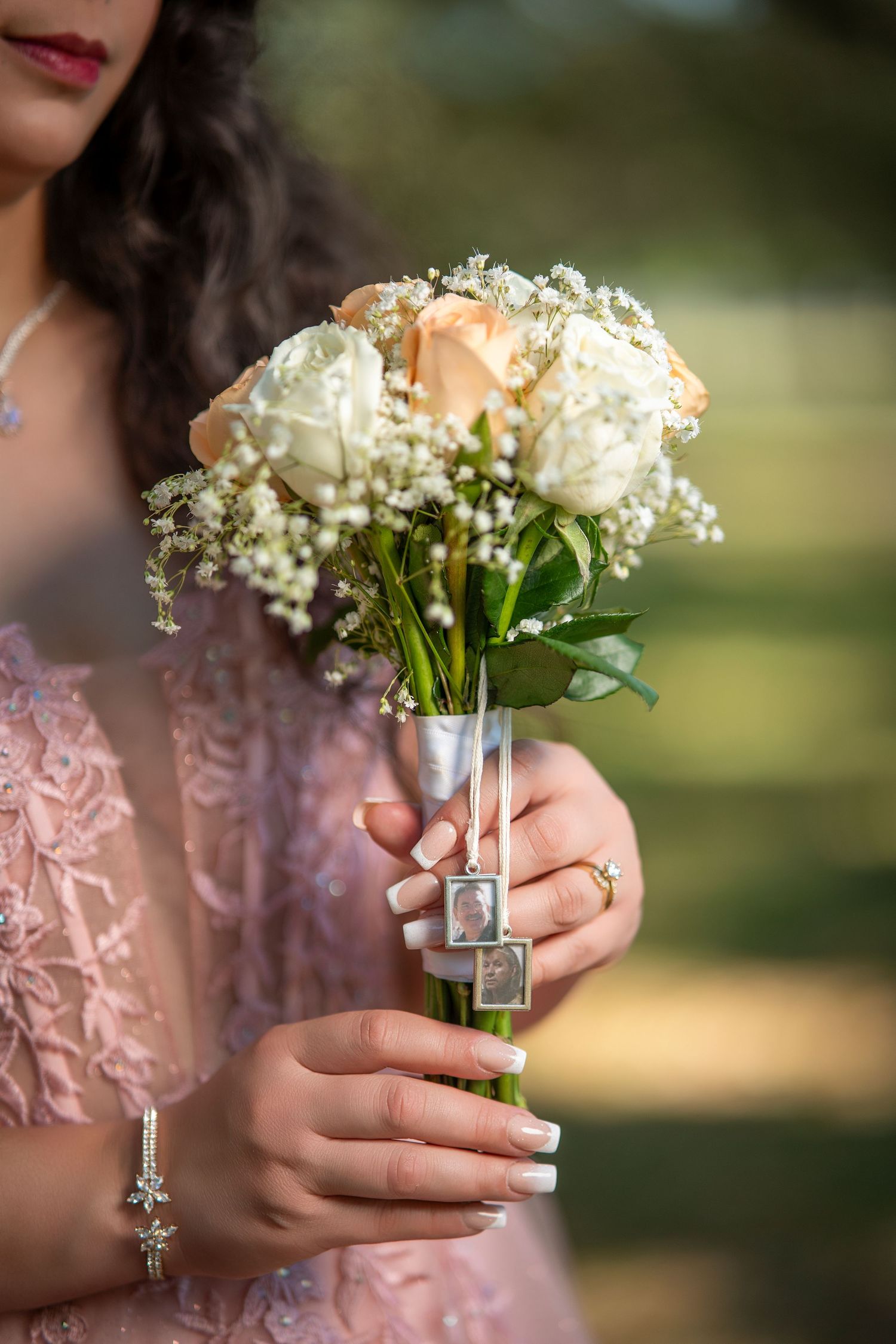 Bride holding a white and peach rose wedding bouquet with memorial photo charms and ribbon, wearing a pink lace dress.