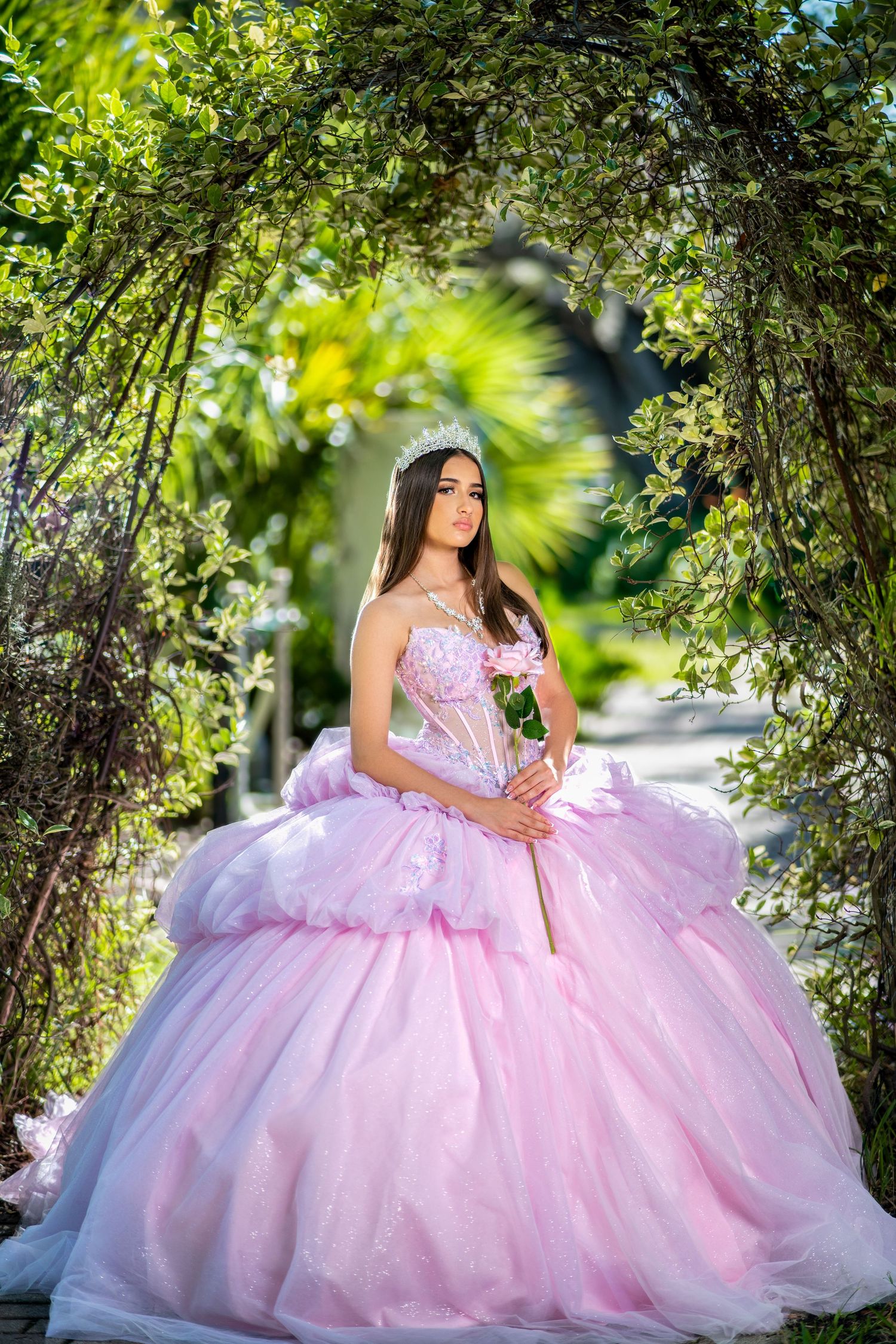 A person in a flowing pink quinceañera gown poses elegantly among lush green foliage in an outdoor garden setting.