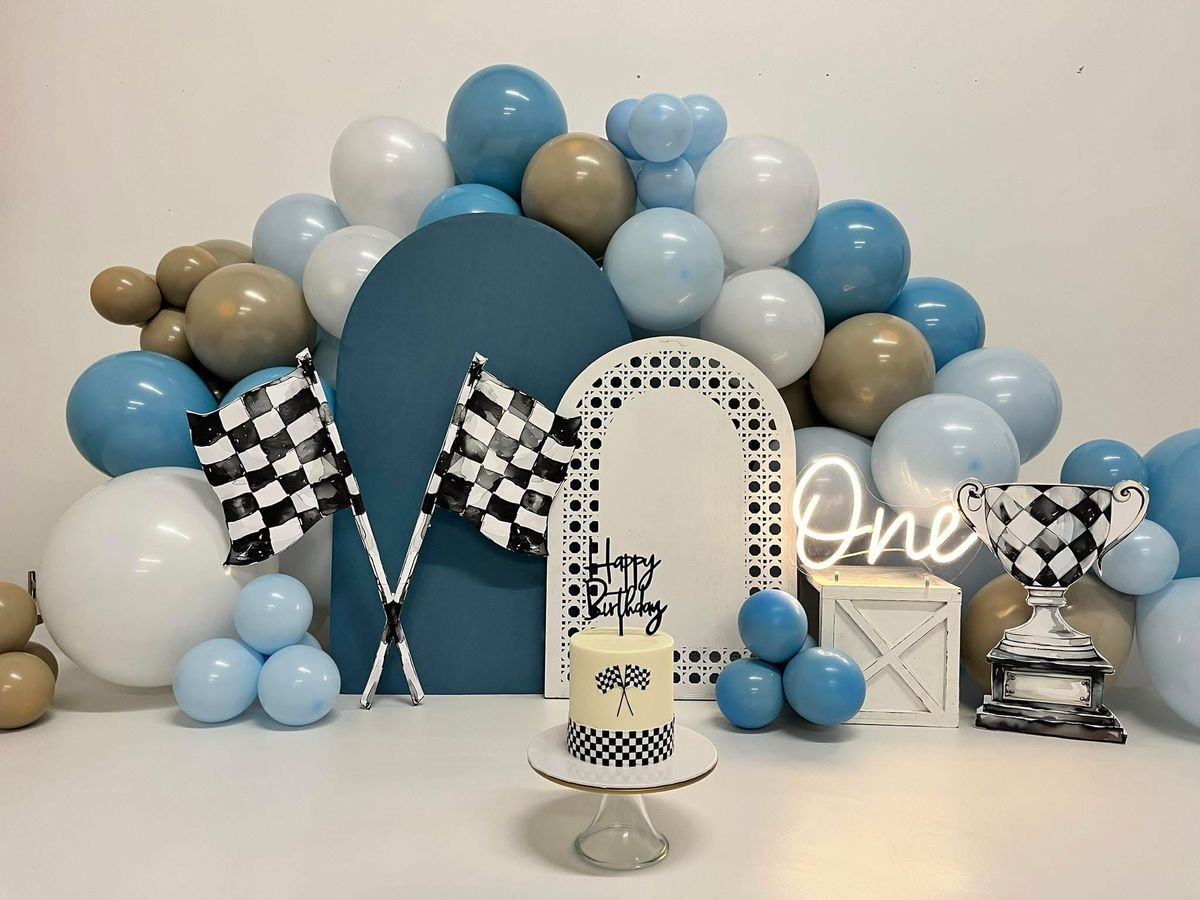 Blue and white balloon arch with racing theme decorations and checkered pattern accents for first birthday party.