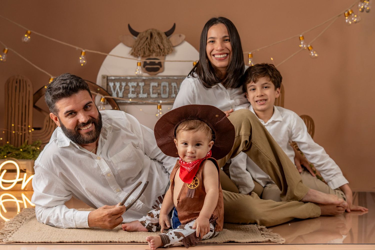 A family in matching white outfits lies together on a beige carpet during a cozy indoor photoshoot.