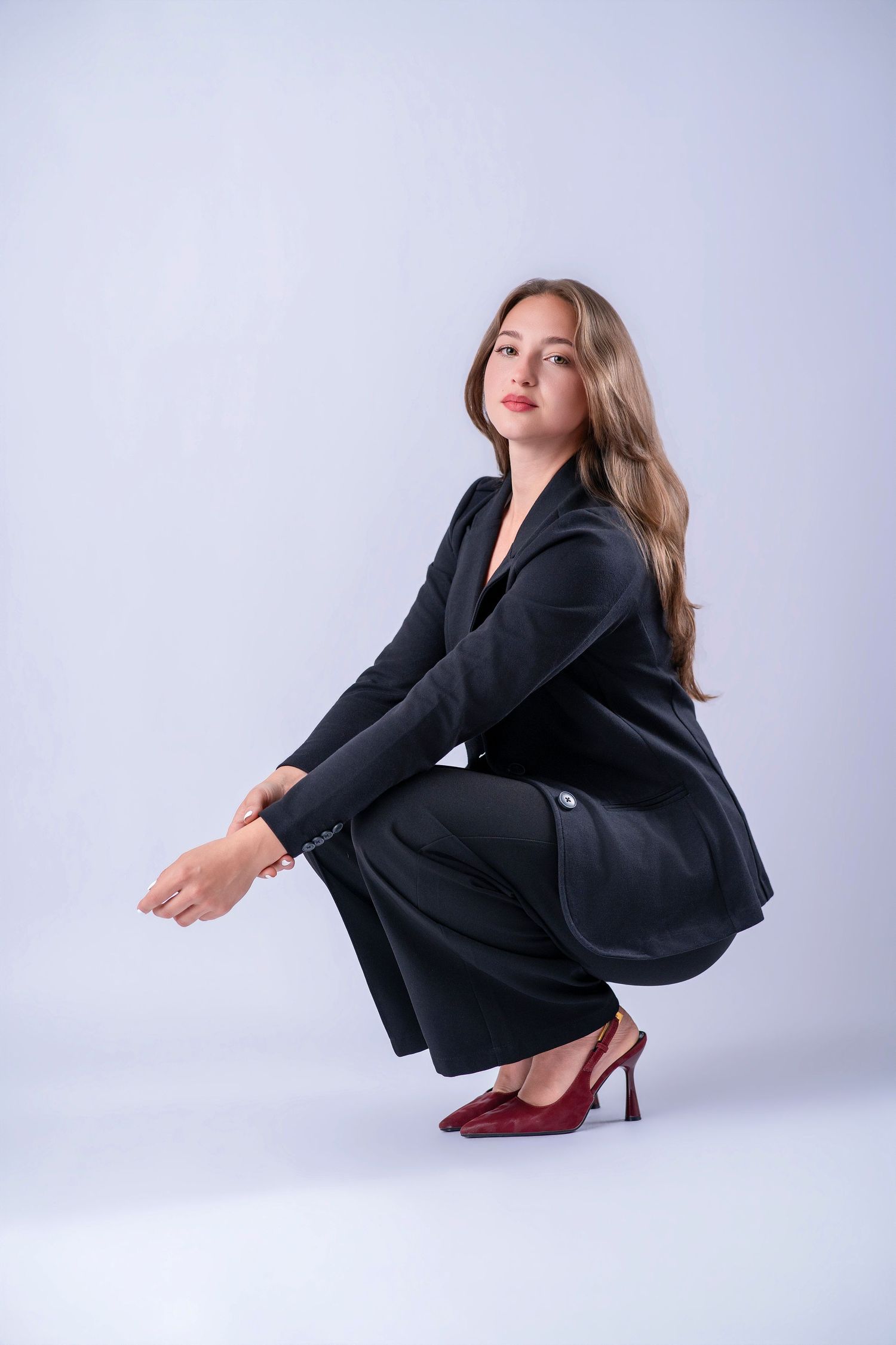 Young woman in black suit and red heels crouching gracefully against a light gray studio background.