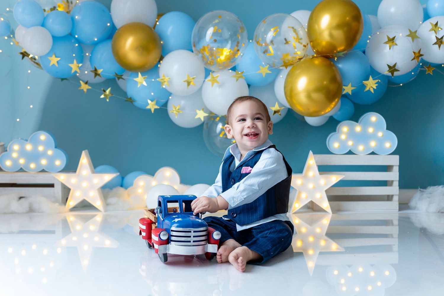 Smiling baby boy in blue vest sits with toy truck at first birthday party with gold and blue balloon arch.