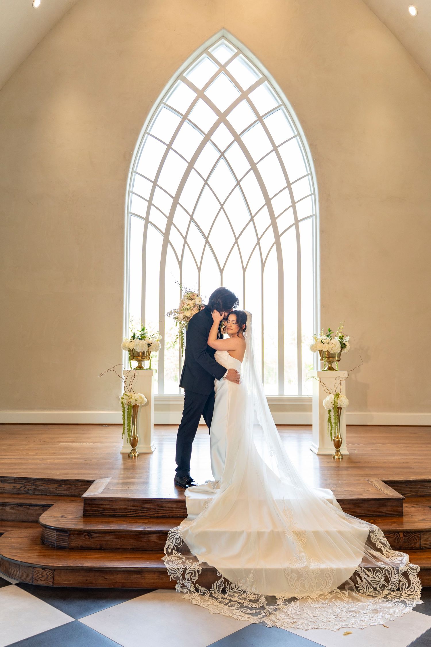 Bride and groom share kiss at altar beneath Gothic arch window in elegant white chapel.