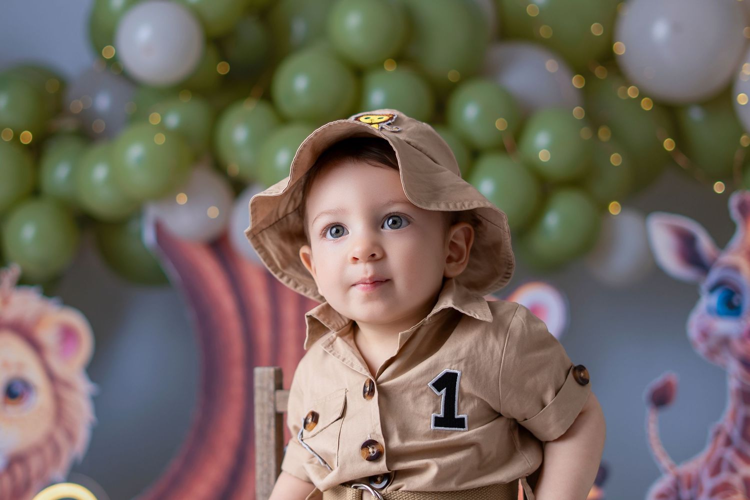 Baby in safari outfit and hat sits before green balloons and jungle animal decorations for first birthday.