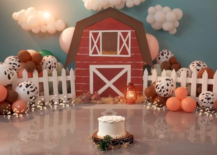 Red barn backdrop with pumpkins and cow print balloons for a rustic first birthday party setup.