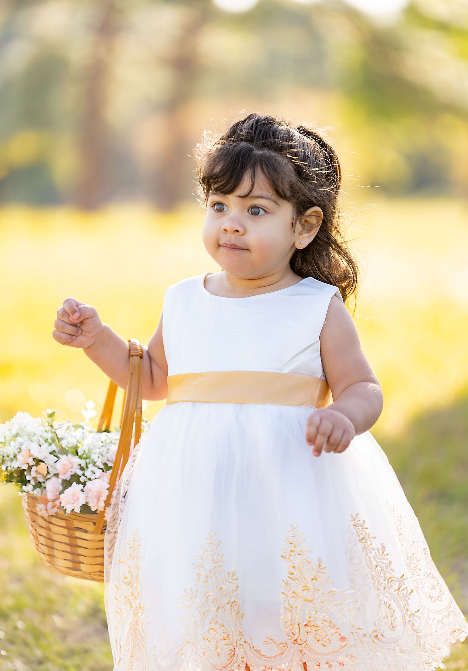 A child in a white dress holding a wicker basket stands in a sunlit meadow with soft golden bokeh background.
