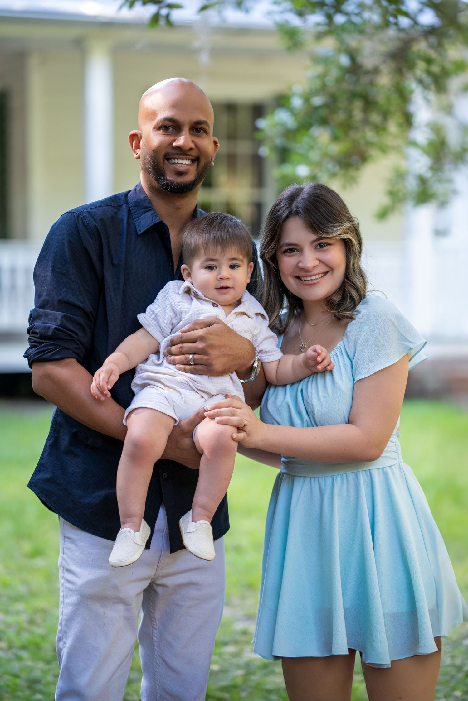 A family of three wearing blue and navy poses outdoors for casual portraits in a backyard setting.