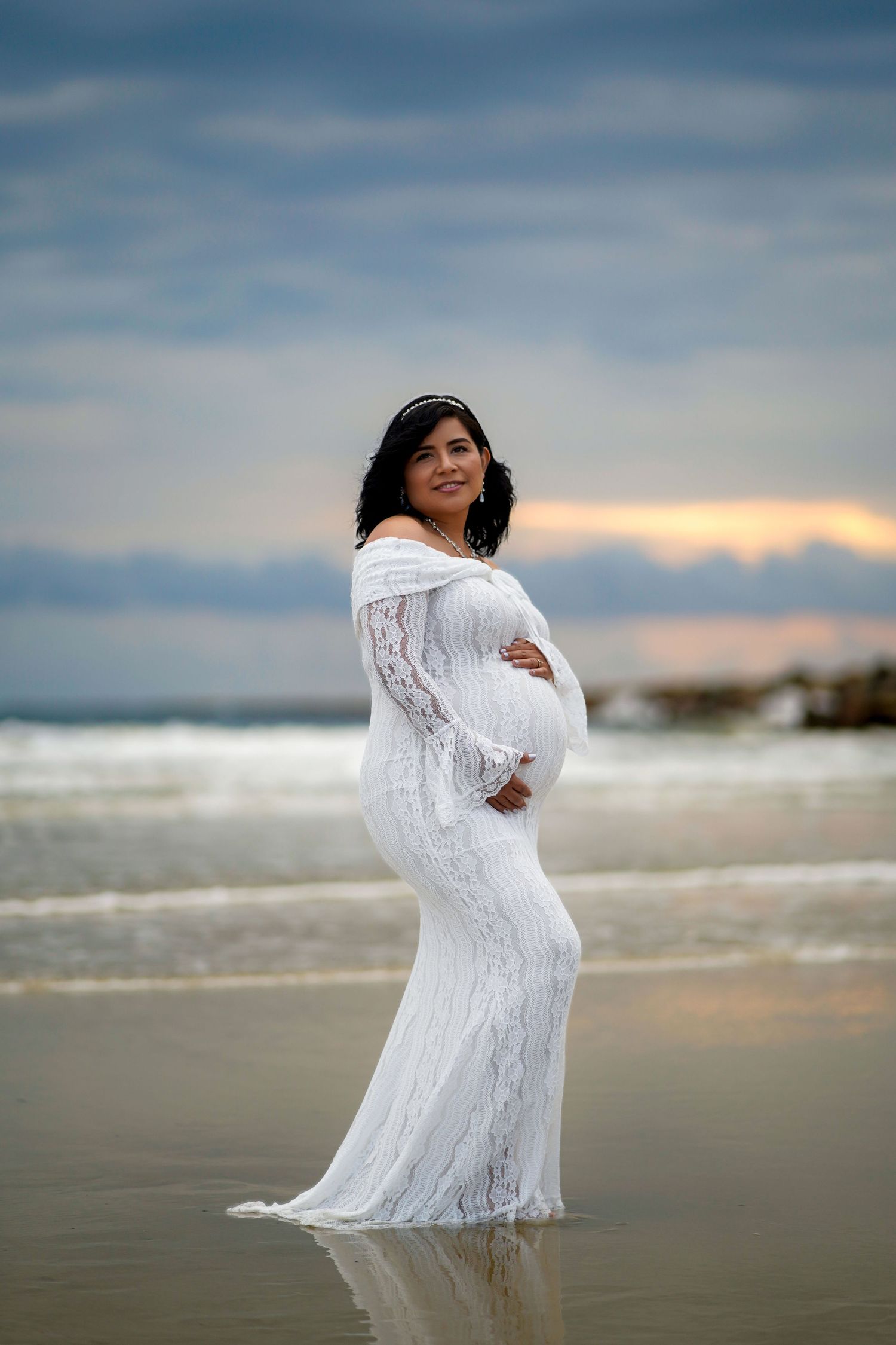A series of photographs showing a white lace maternity dress photoshoot on a cloudy beach at sunset.