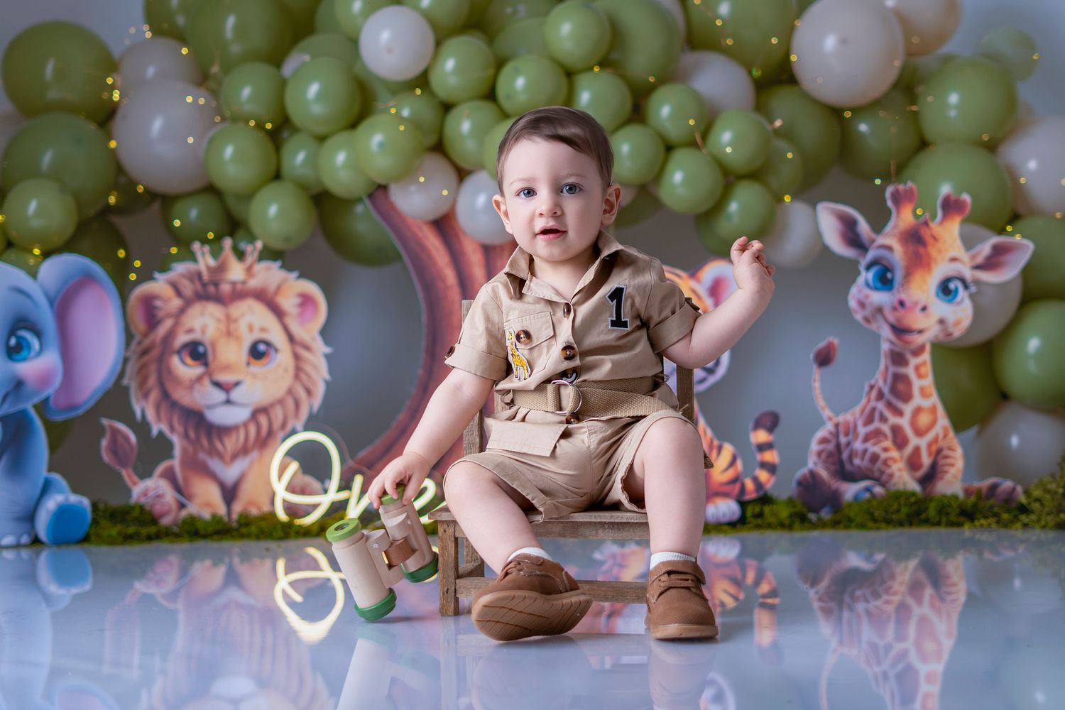 Baby boy in safari outfit sits before jungle animal backdrop with green balloons at first birthday cake smash photo session.