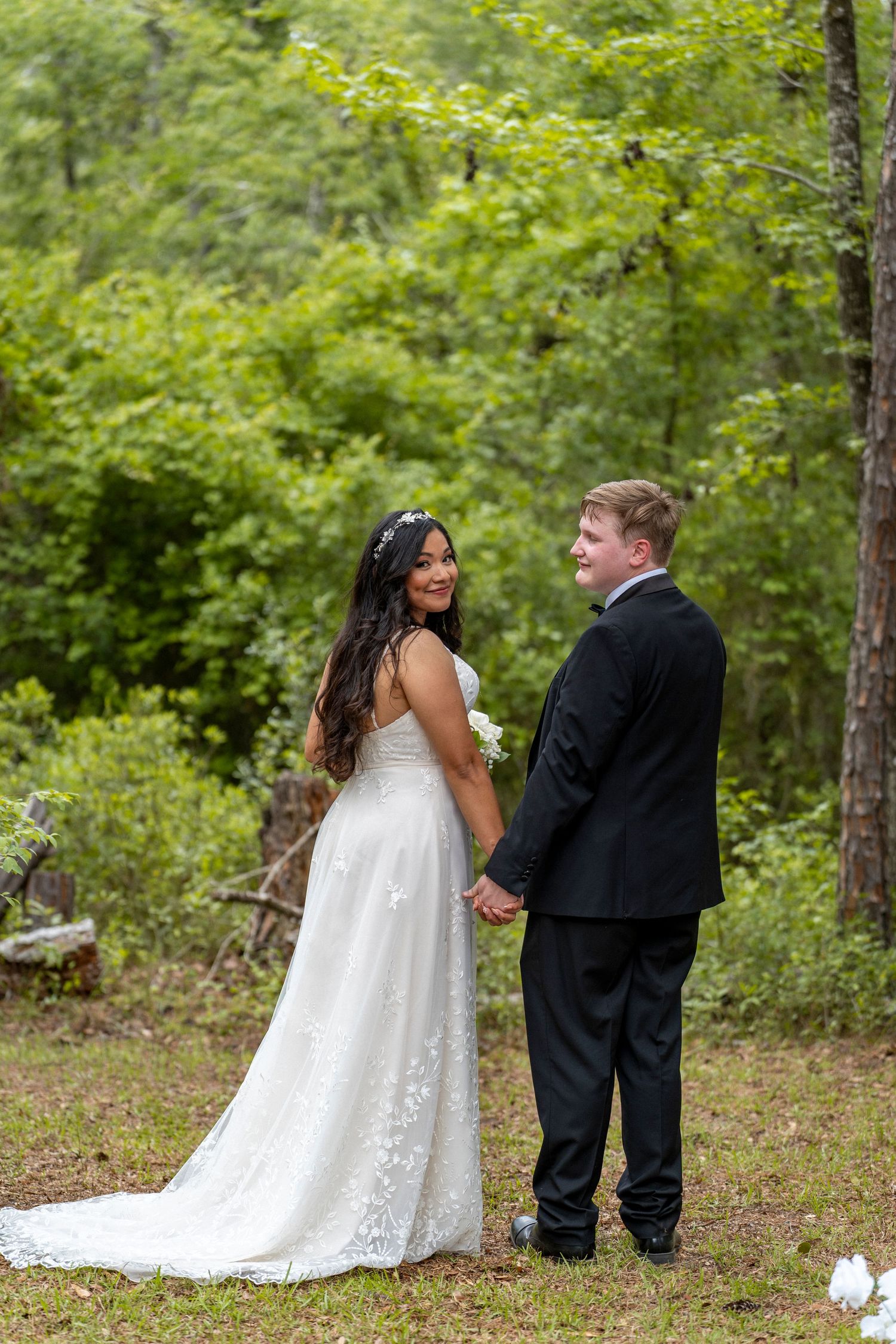 A bride in a white lace gown and groom in a black tuxedo hold hands while posing in a lush green forest setting.