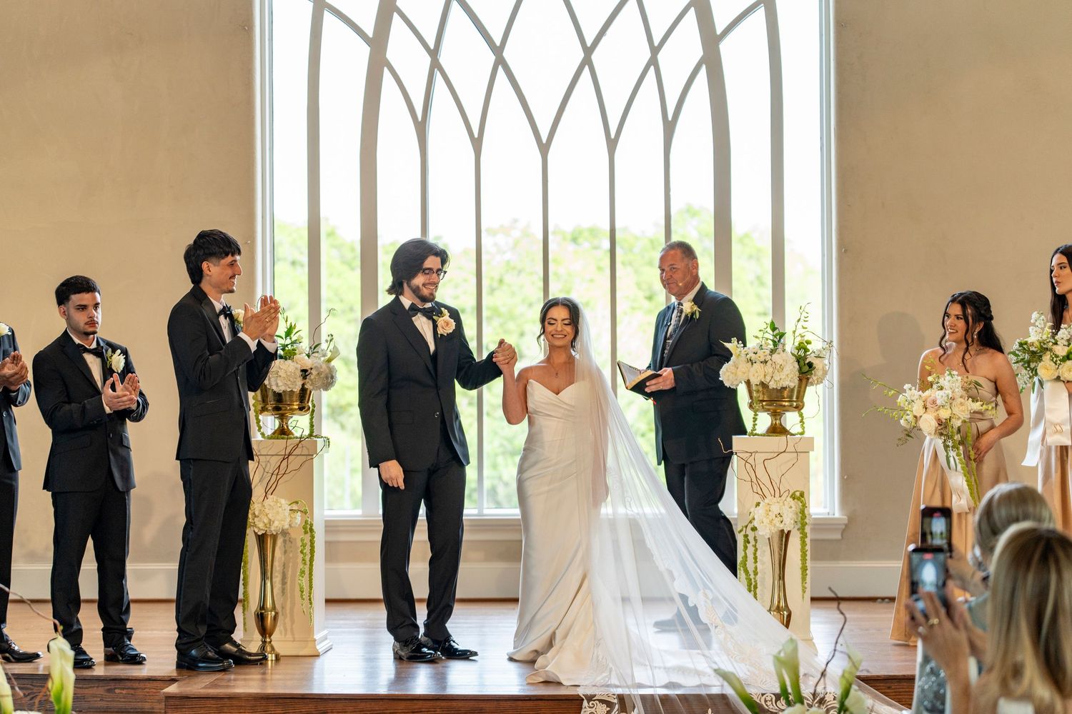 Bride and groom celebrate at wedding altar with bridal party in elegant chapel with arched windows.