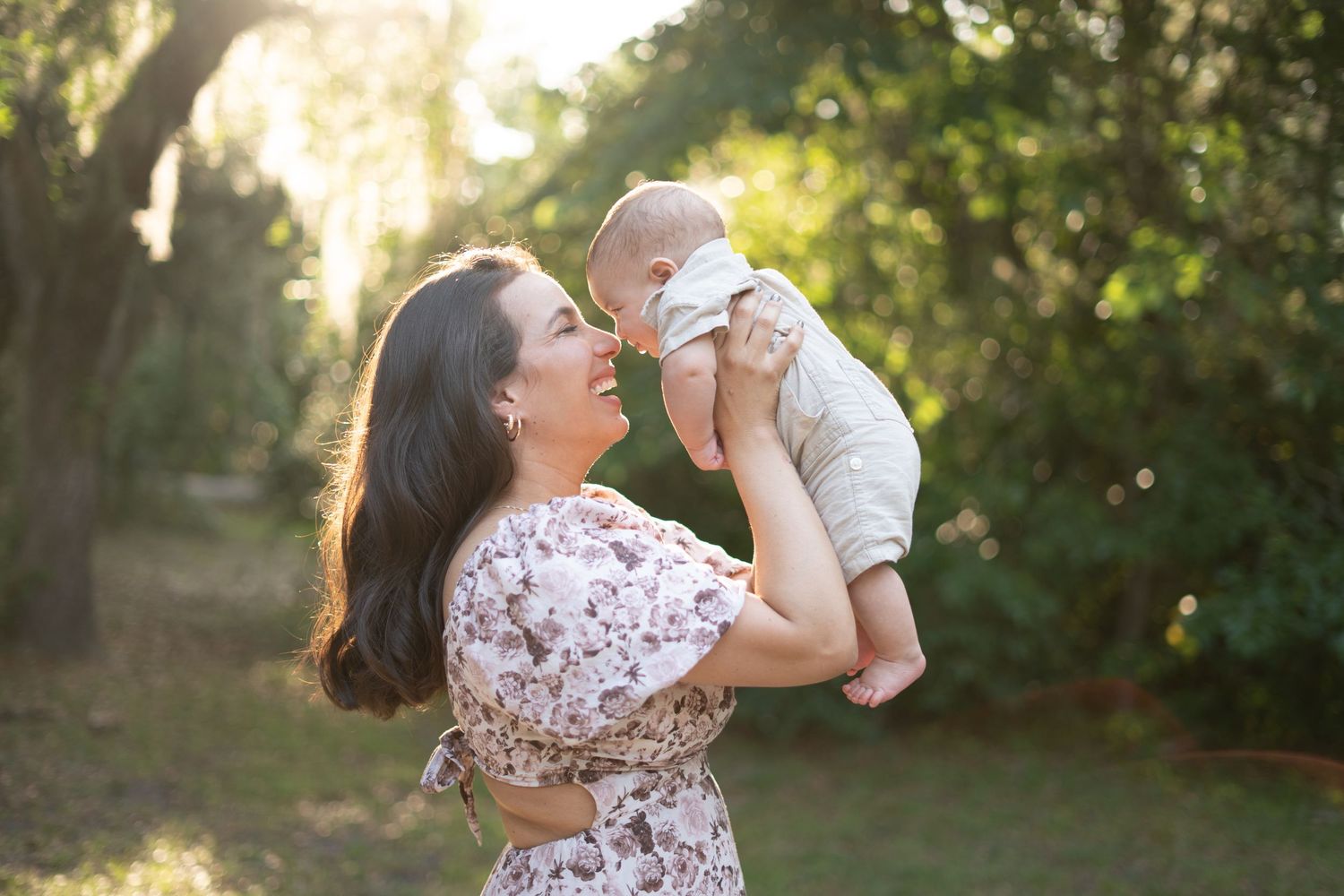 Mother in floral dress lifts small baby in white onesie during golden hour outdoor photoshoot.