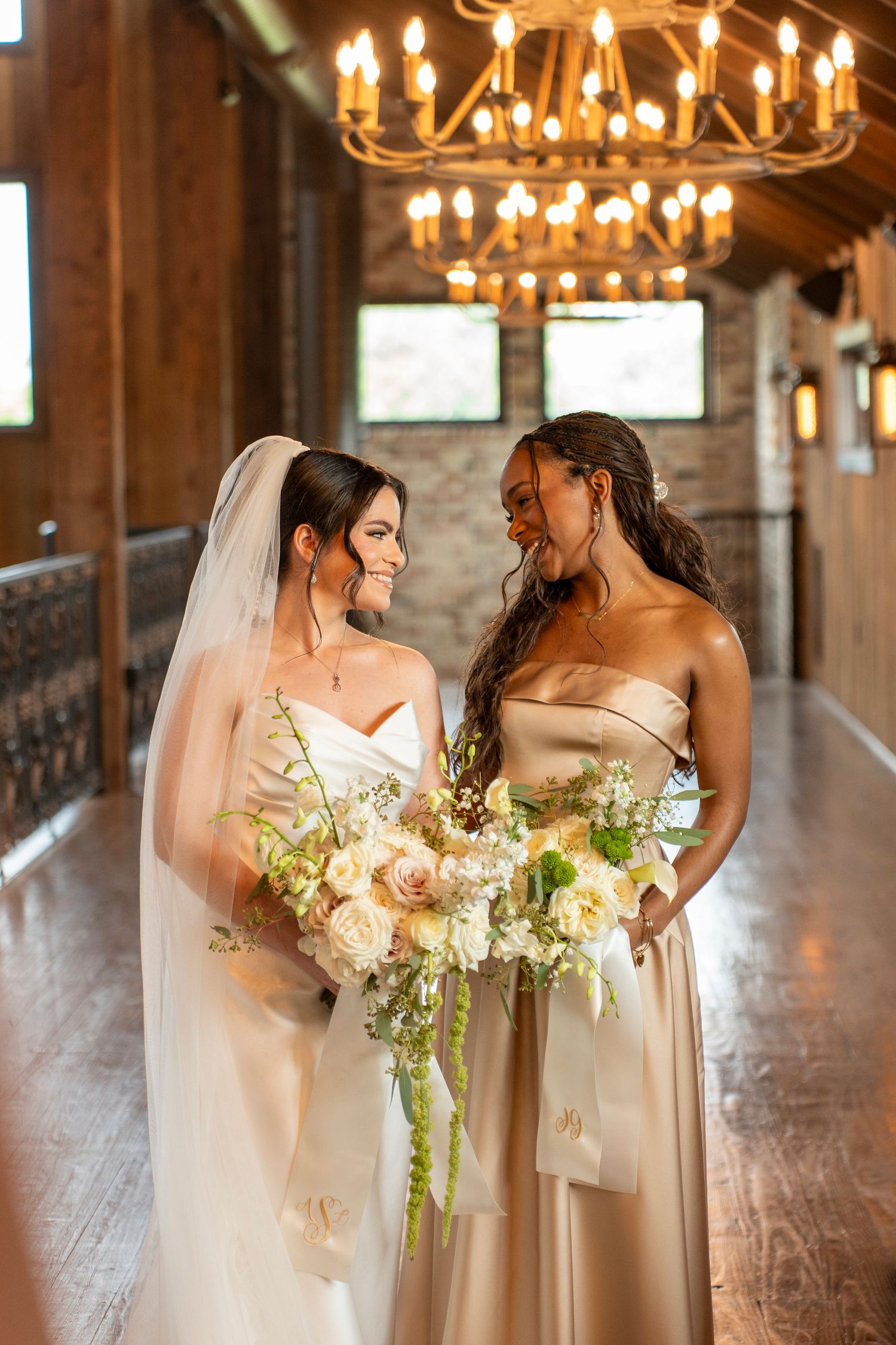 Bride in white gown with veil holds floral bouquet beside bridesmaid in champagne dress in elegant venue.
