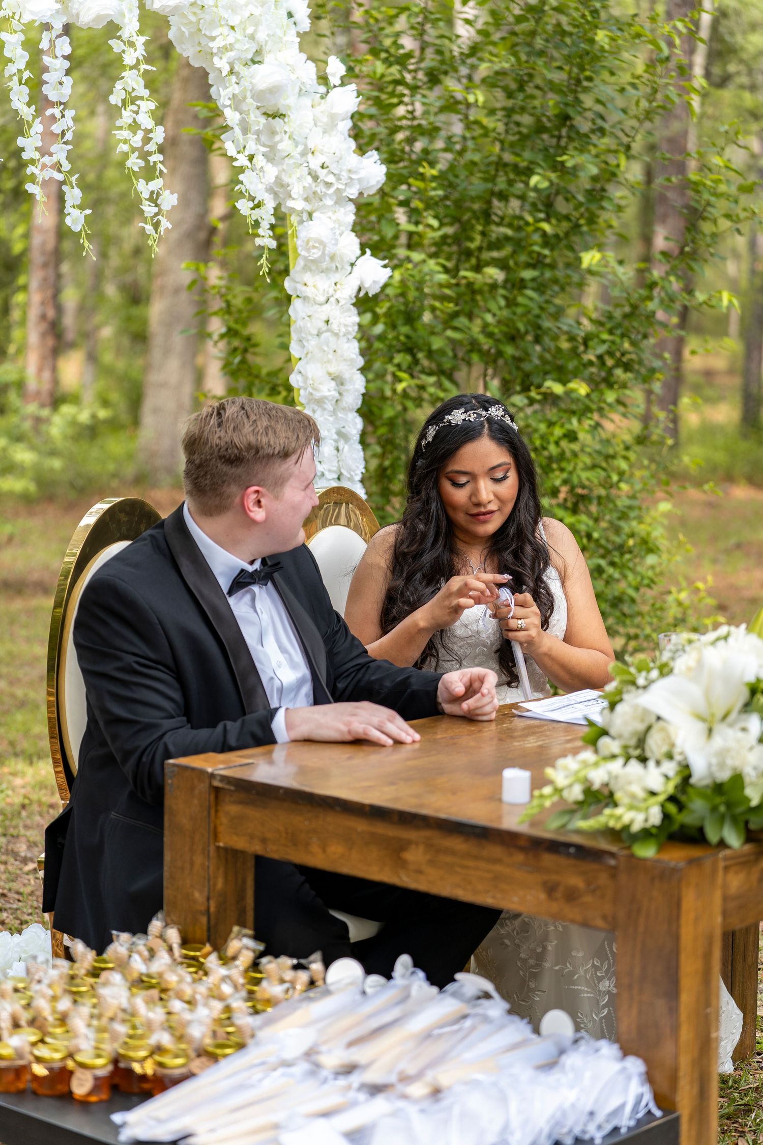 Bride and groom signing wedding documents at a wooden table outdoors, surrounded by white flowers and greenery.