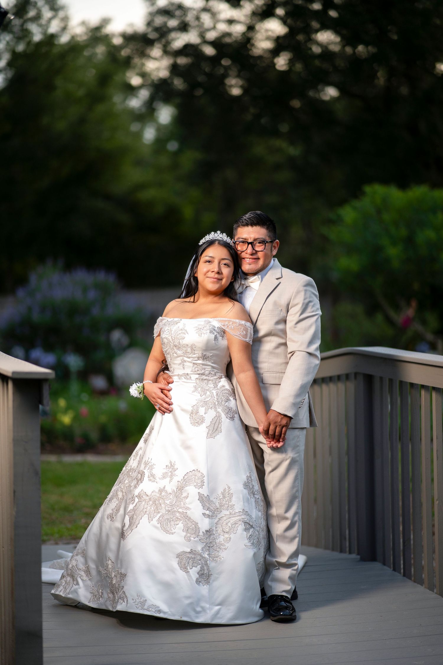 Newlyweds pose on a garden bridge, bride in white gown with tiara, groom in tan suit, smiling.