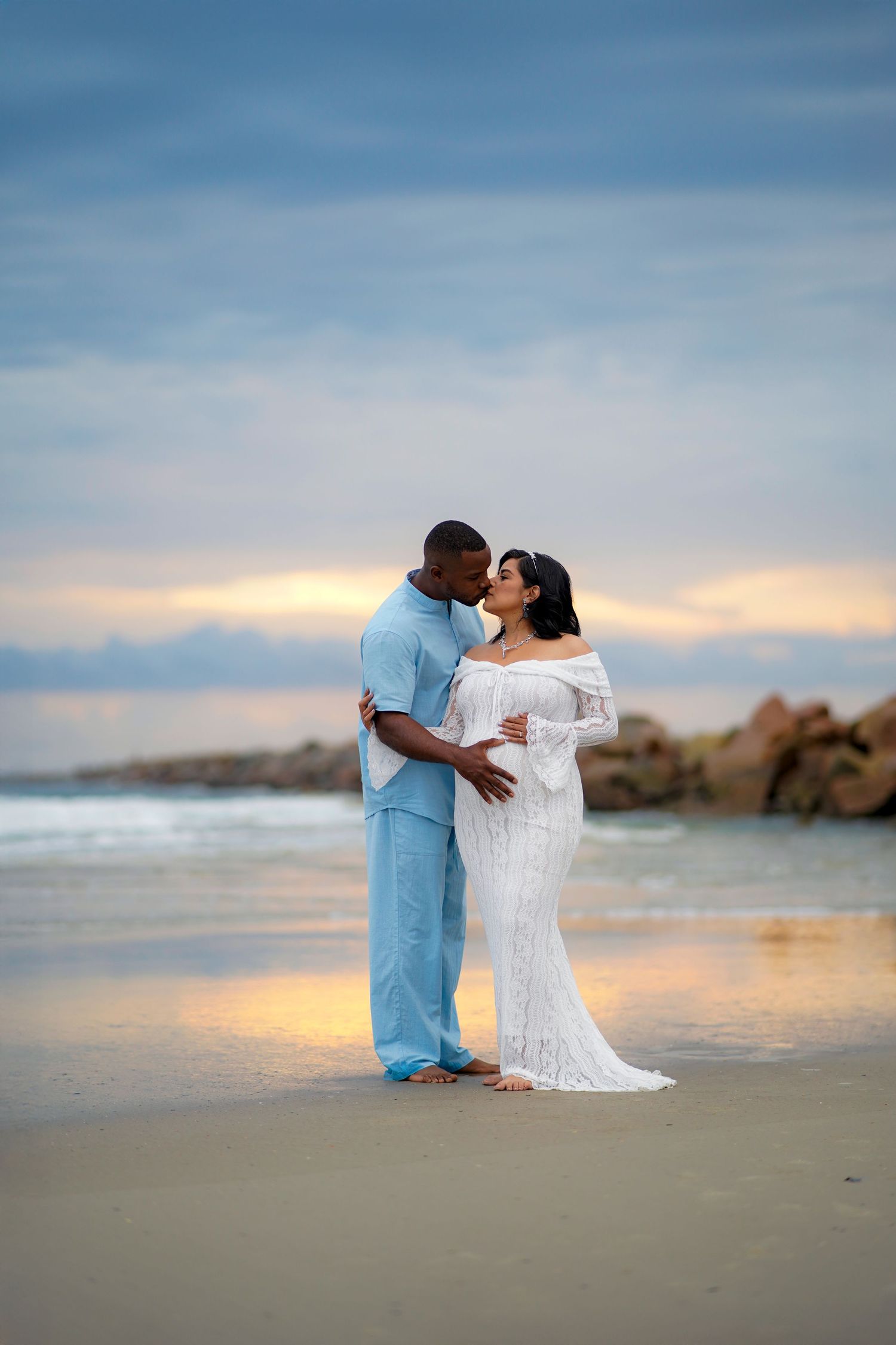 A couple embraces on a sandy beach at sunset with ocean waves and rocky outcrops in the background.
