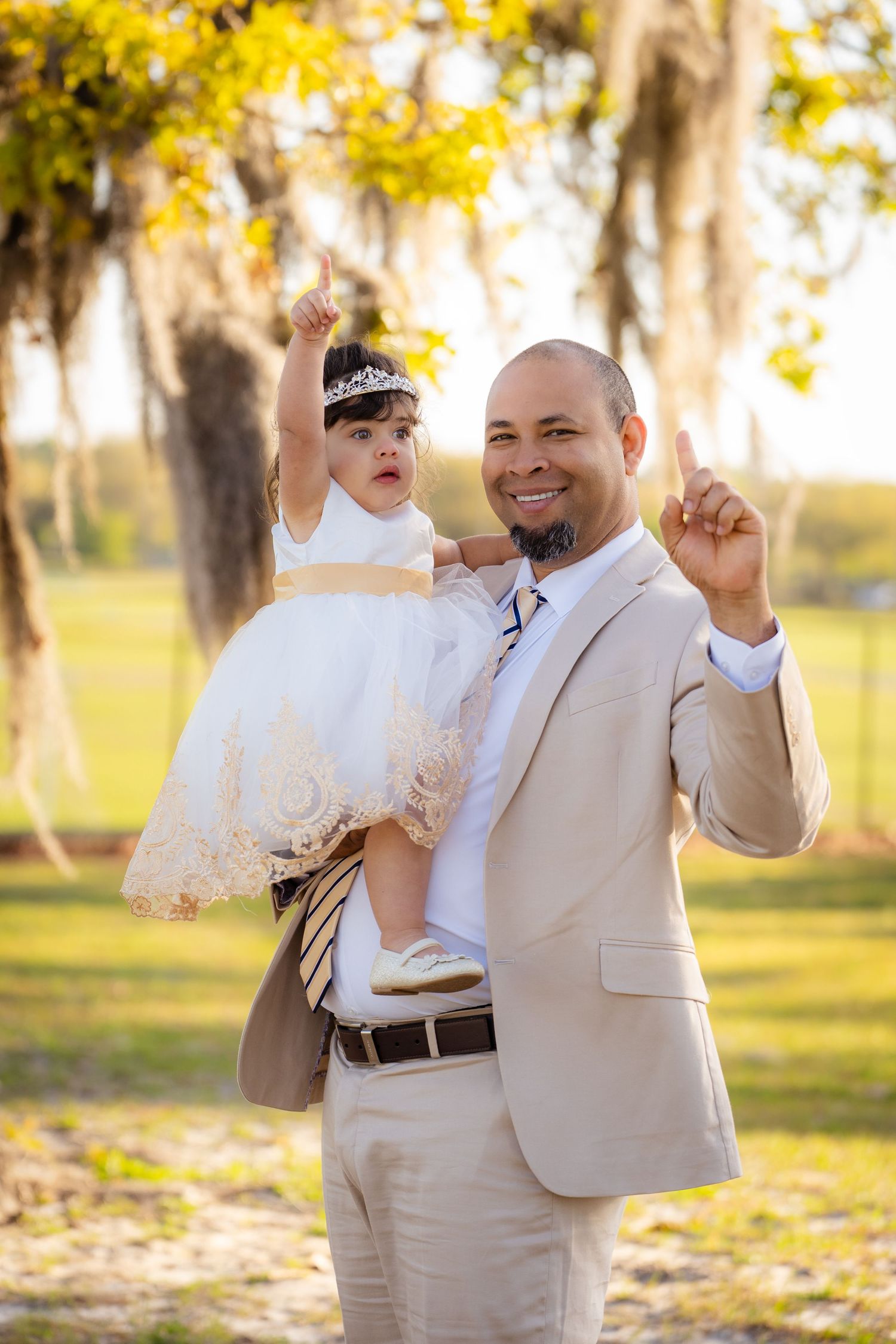 Father and son wearing matching beige suits dance joyfully together in a sunlit park setting.