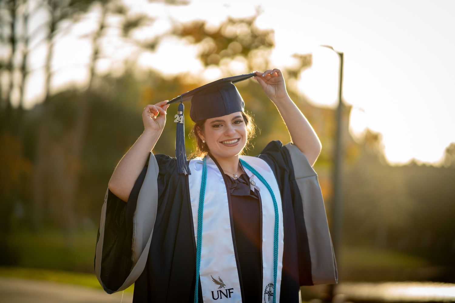 Graduate in black cap and gown with blue stole celebrates during golden hour sunset outdoors.