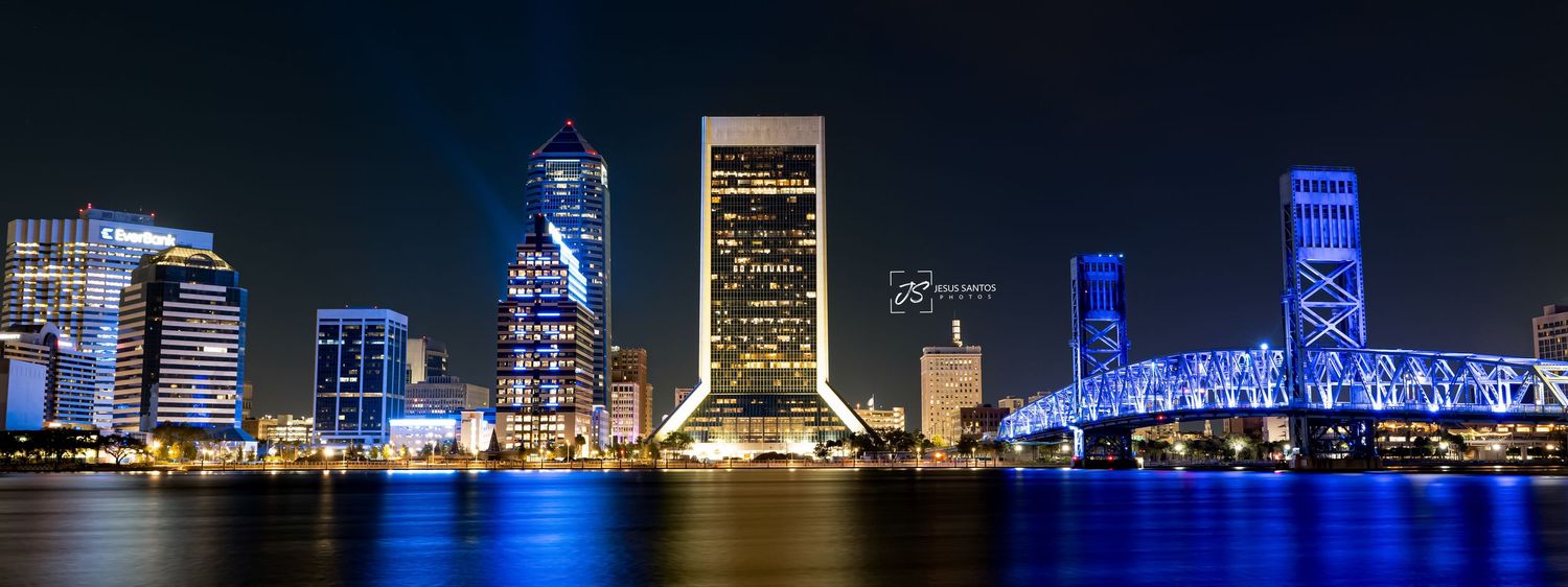 Panoramic night skyline of Jacksonville Florida with illuminated bridges and buildings reflecting on St. Johns River.