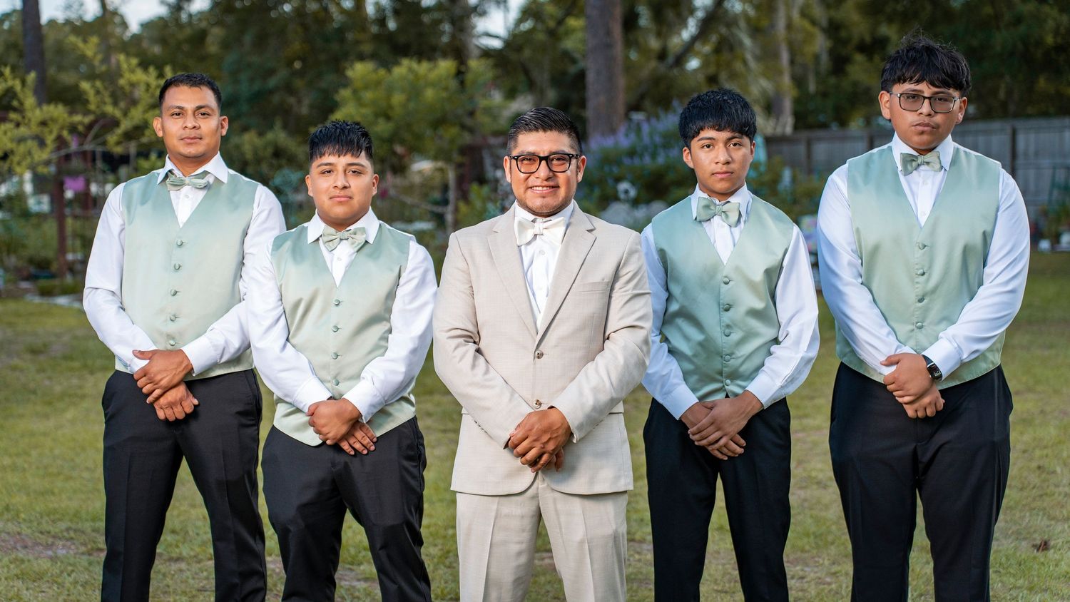 Groom in beige suit with four groomsmen in mint green vests and bow ties posing outdoors at a wedding.