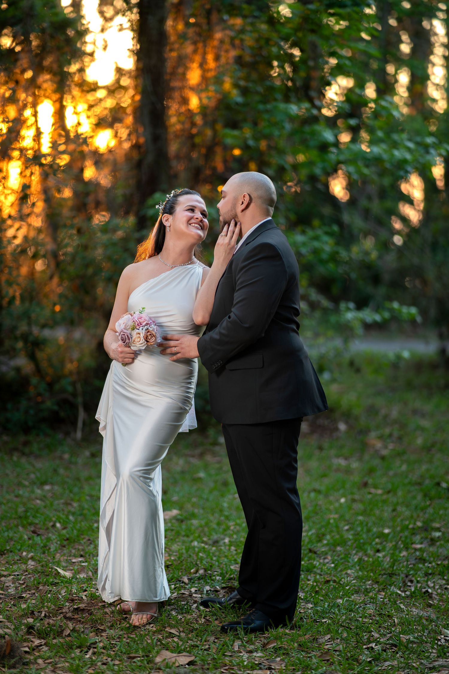 Bride in white gown holds bouquet while groom in black suit tenderly cups her face, sharing a romantic moment at sunset.