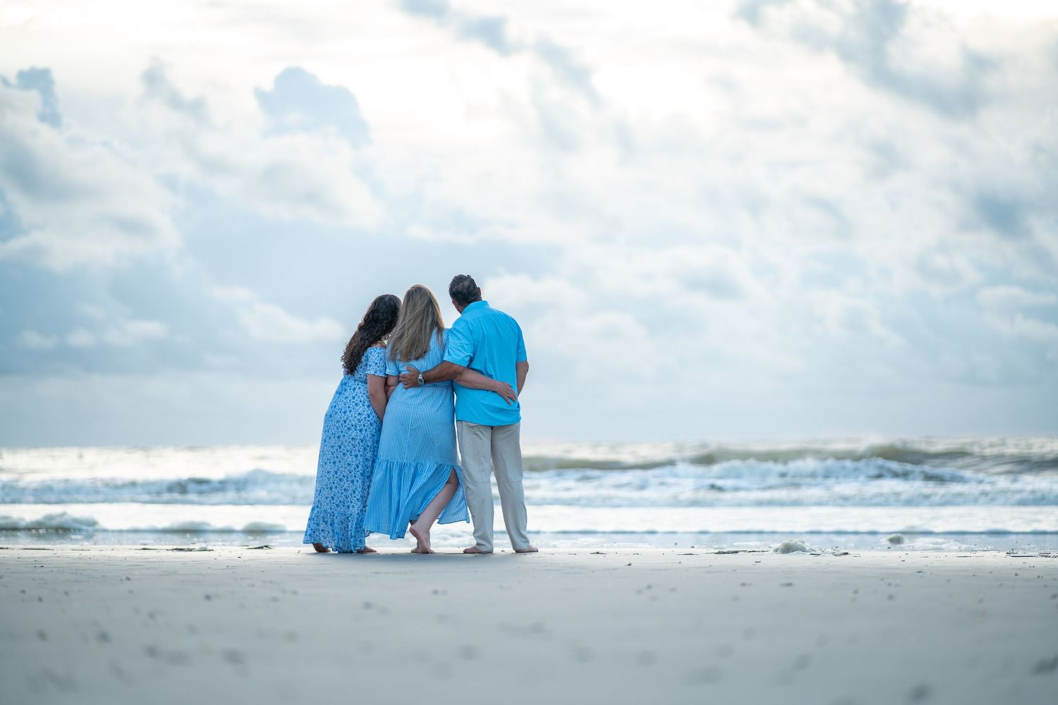 A couple in blue attire embraces on a cloudy beach with crashing waves in the background.