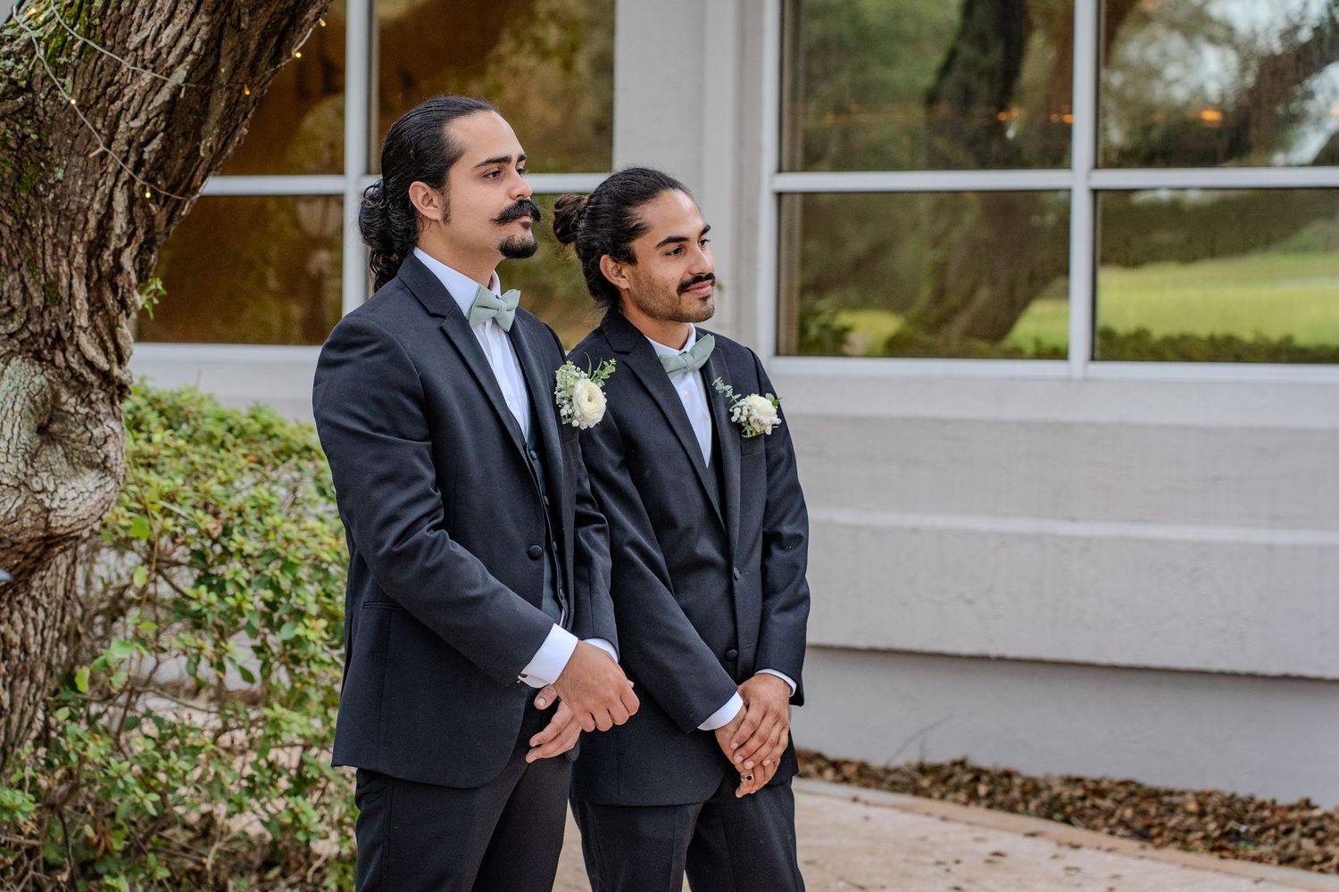 Two people in dark suits stand together outdoors while sharing a friendly moment.