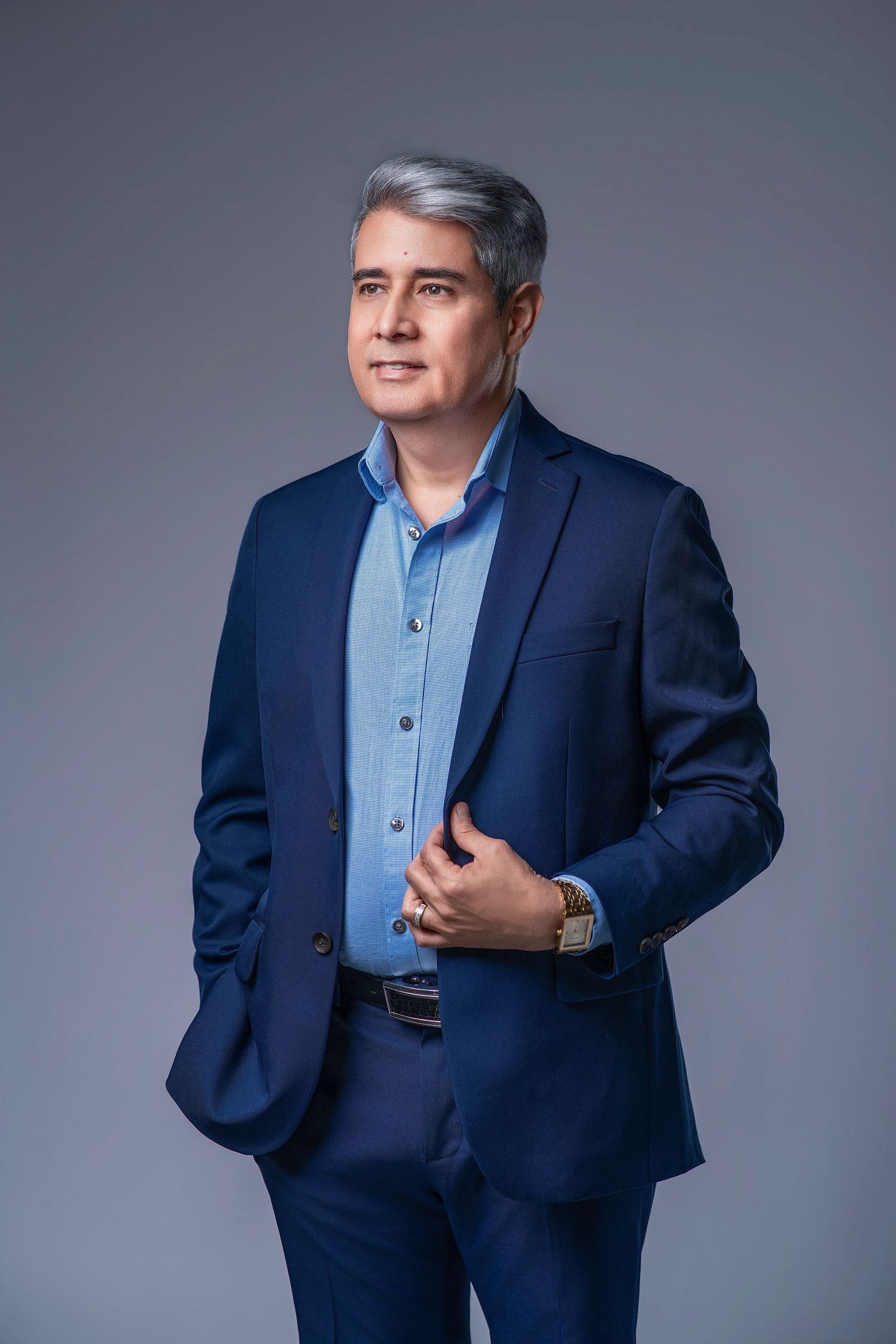 Middle-aged man with gray hair wearing a navy blue suit and light blue shirt, posing confidently against a gray background.