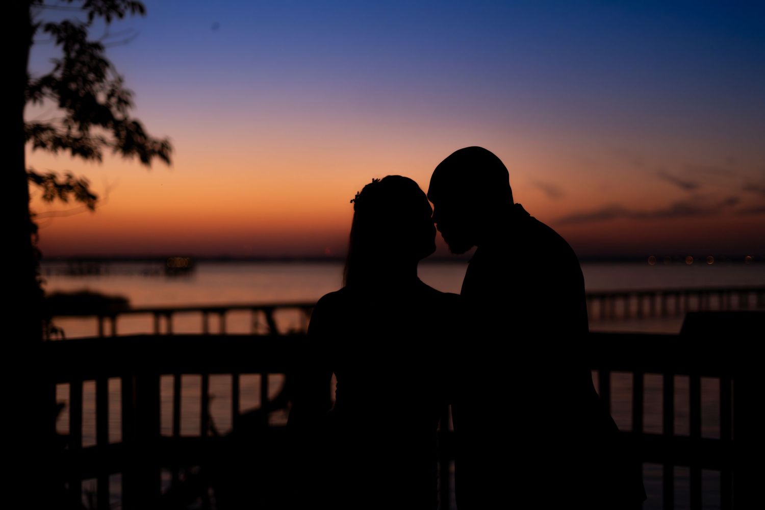 Silhouette of a romantic couple sharing a kiss on a waterfront deck at sunset with vibrant orange and blue sky.
