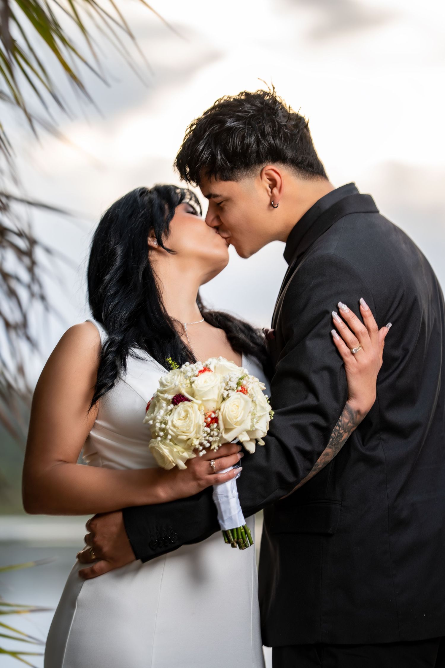 Romantic wedding couple shares intimate kisses in four sequential photos holding white rose bouquet against palm trees.