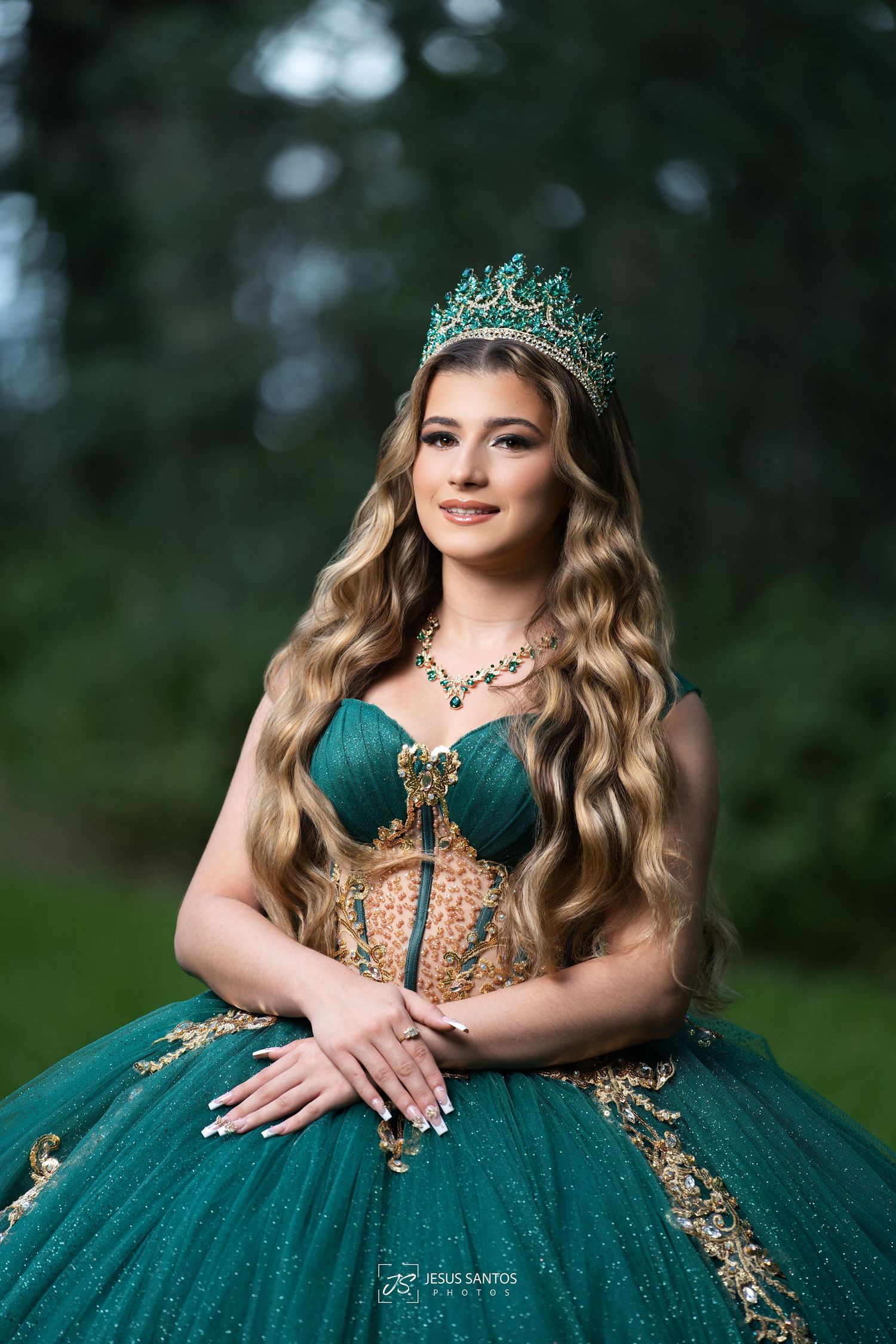 Young woman in emerald green quinceañera ball gown with gold accents, wearing a jeweled crown and long wavy hair outdoors.