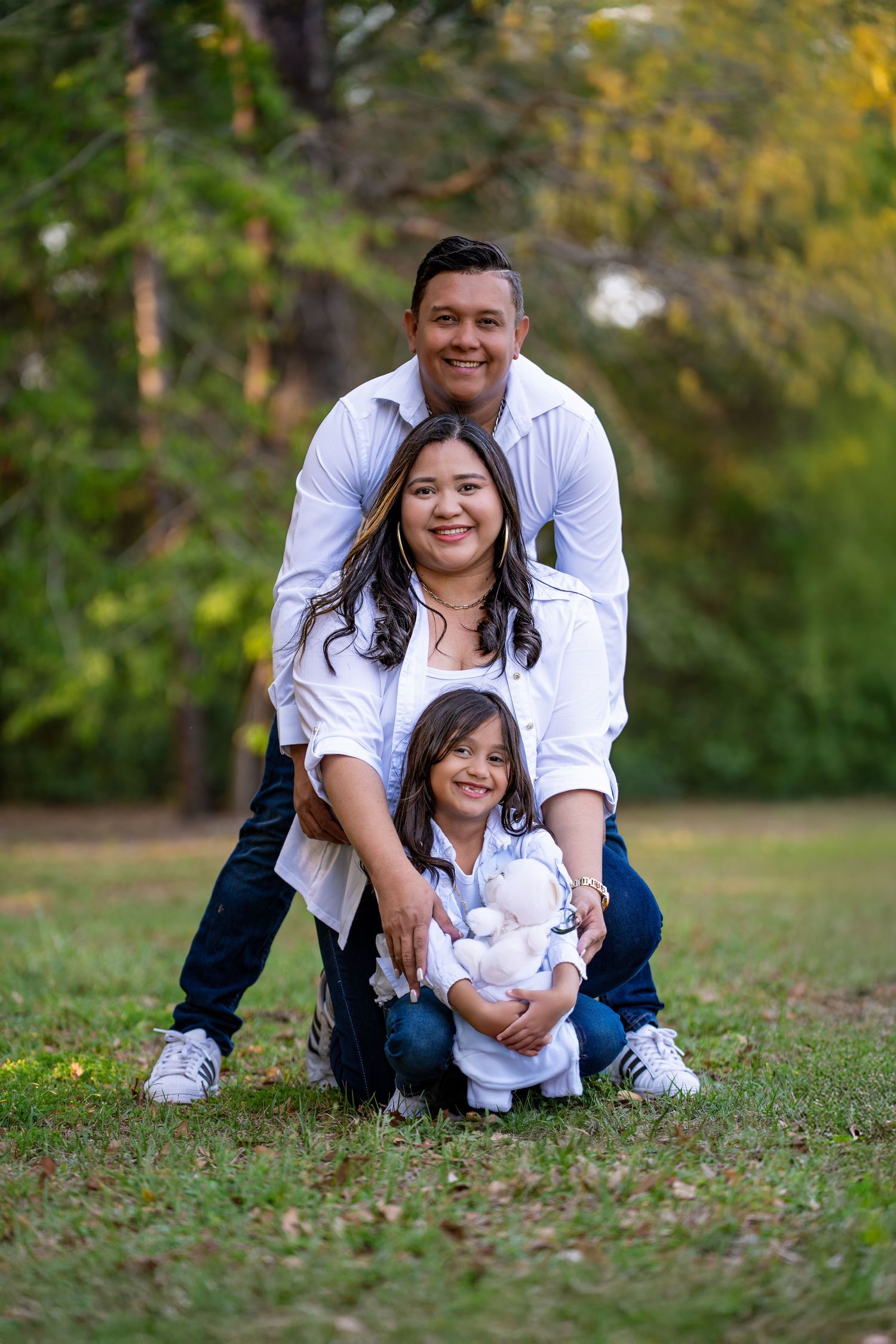 A family wearing coordinated white outfits poses together outdoors in a park with autumn trees in the background.
