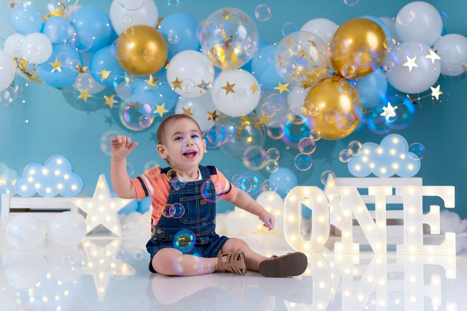 Baby boy celebrates first birthday with gold and blue balloon backdrop and illuminated ONE sign.