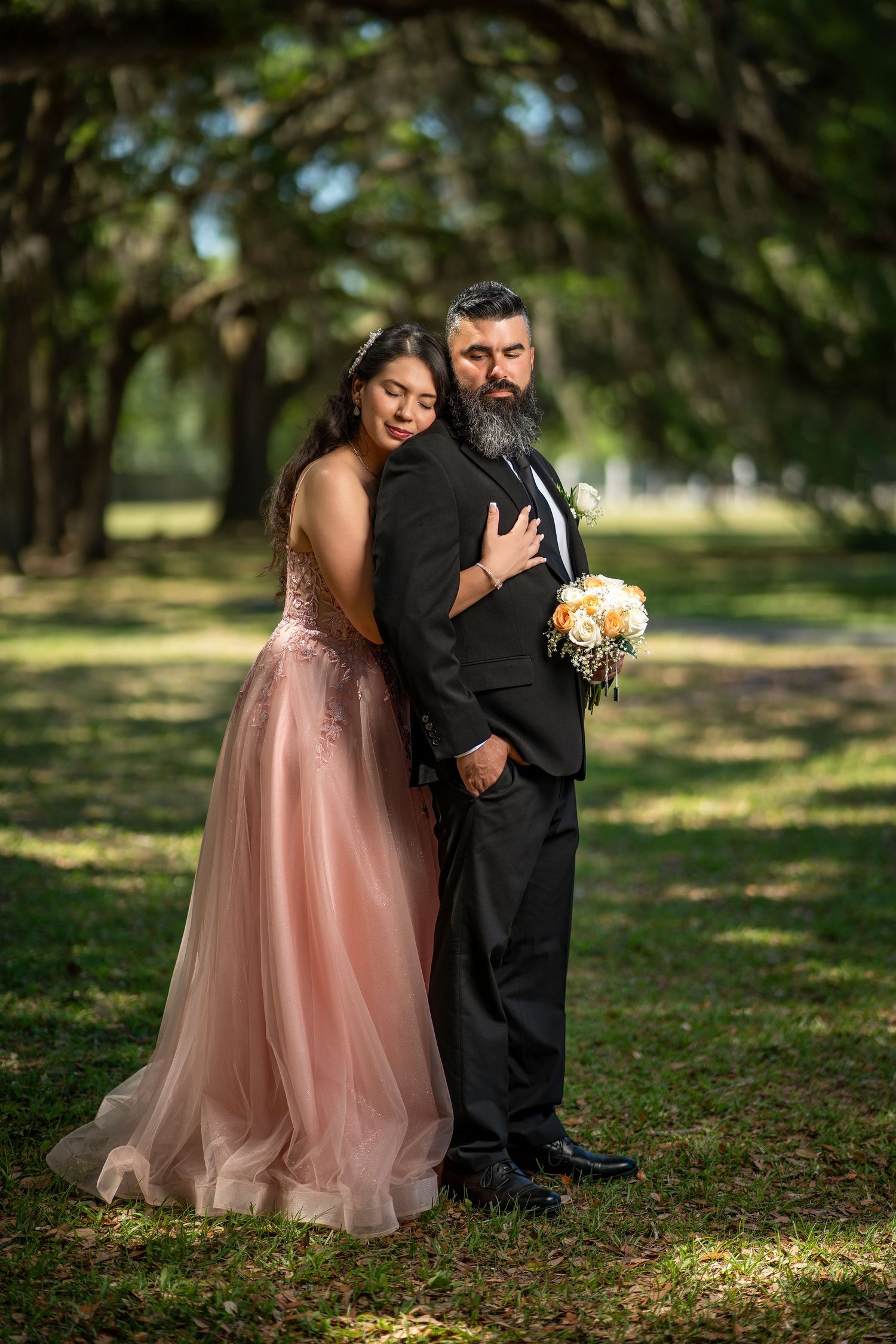 A bearded groom in a black tuxedo embraces a young woman in a blush pink gown holding a bouquet outdoors.