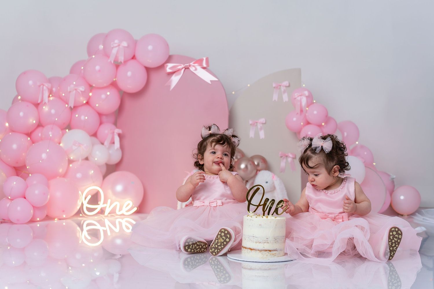Pink birthday celebration setup with balloon arch, neon lights, and tulle dresses for a first birthday cake smash photo session.