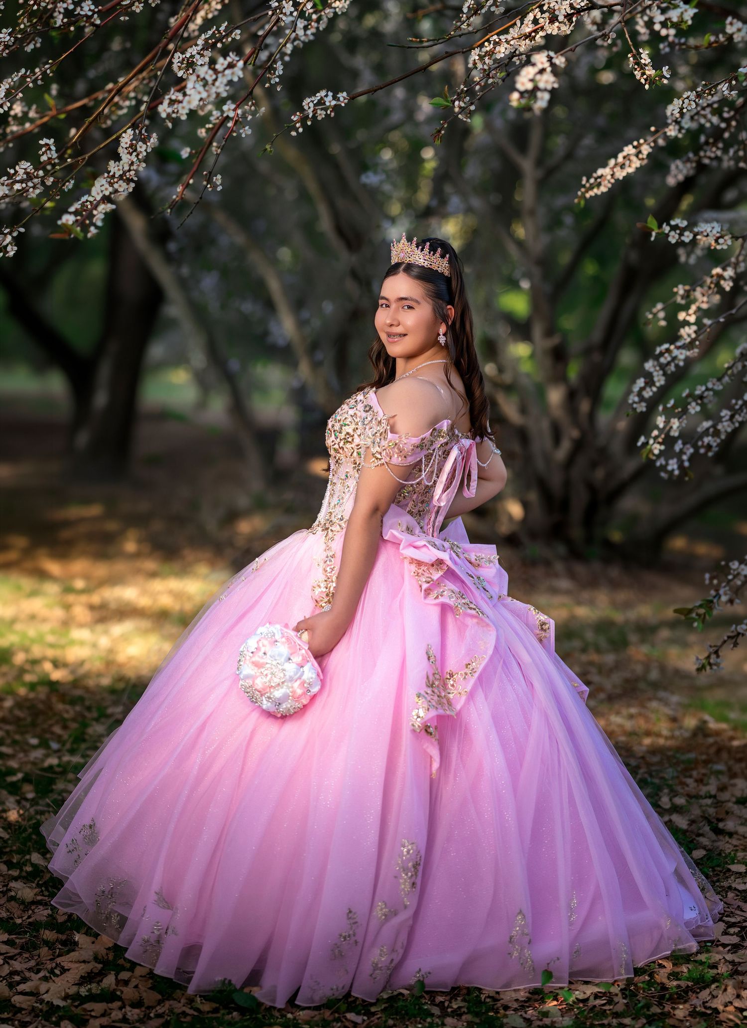 Young woman in pink quinceañera ball gown with gold embroidery and tiara poses among blooming cherry blossom trees.