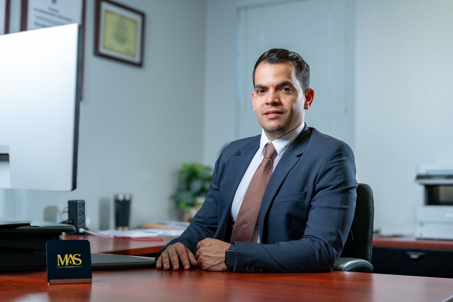 Professional wearing a dark suit sits at a wooden desk in a modern office setting.