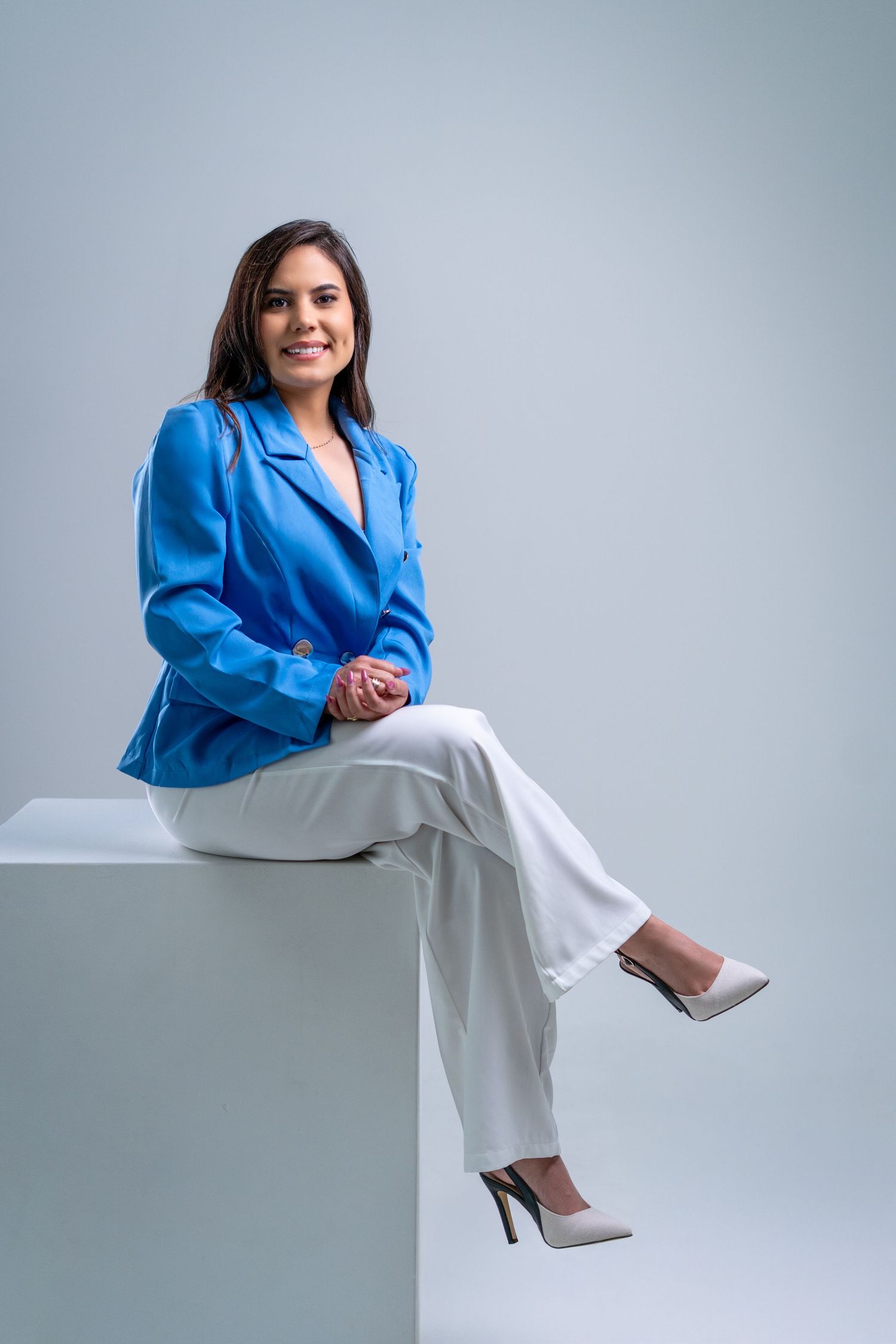 Professional studio portraits of someone wearing a bright blue blazer and white pants seated against a light background.