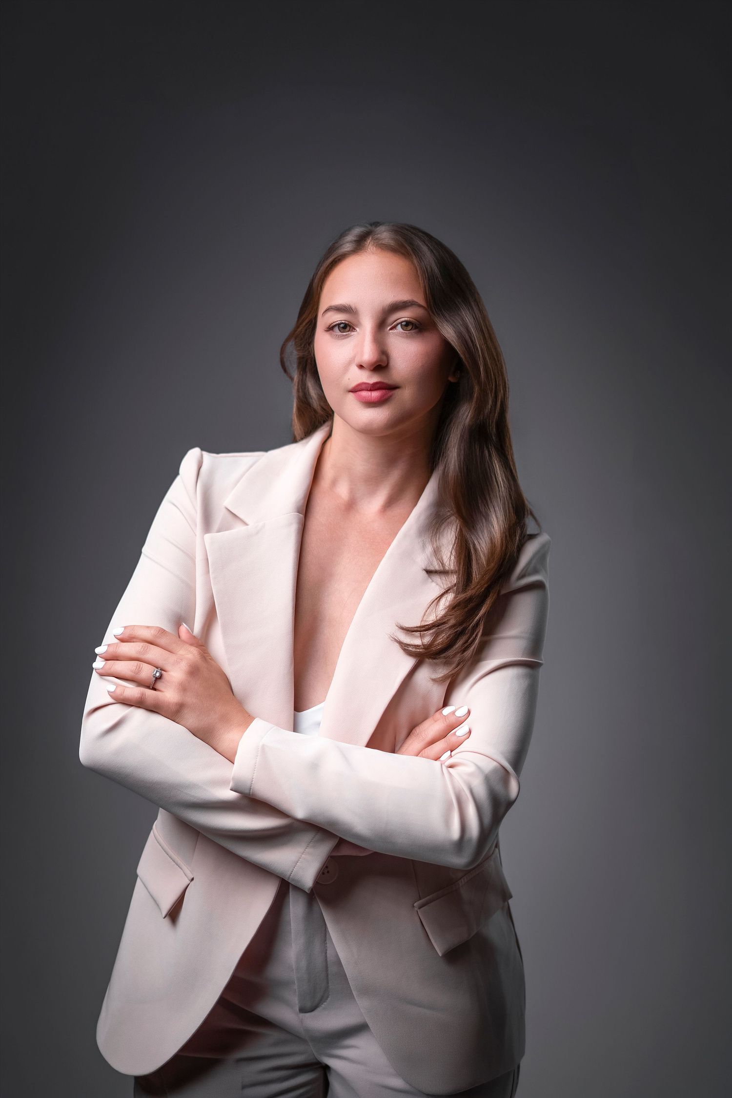 Young woman in white blazer suit with arms crossed, posing confidently against gray studio background.