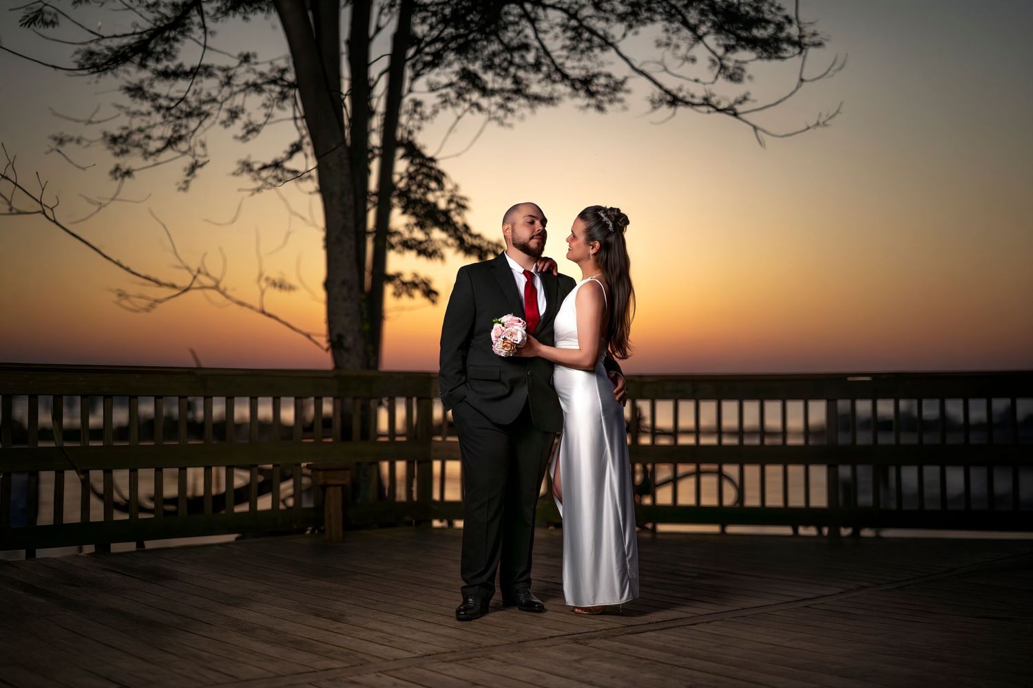 Bride and groom share a romantic moment on a wooden deck at sunset, with a serene waterfront backdrop.