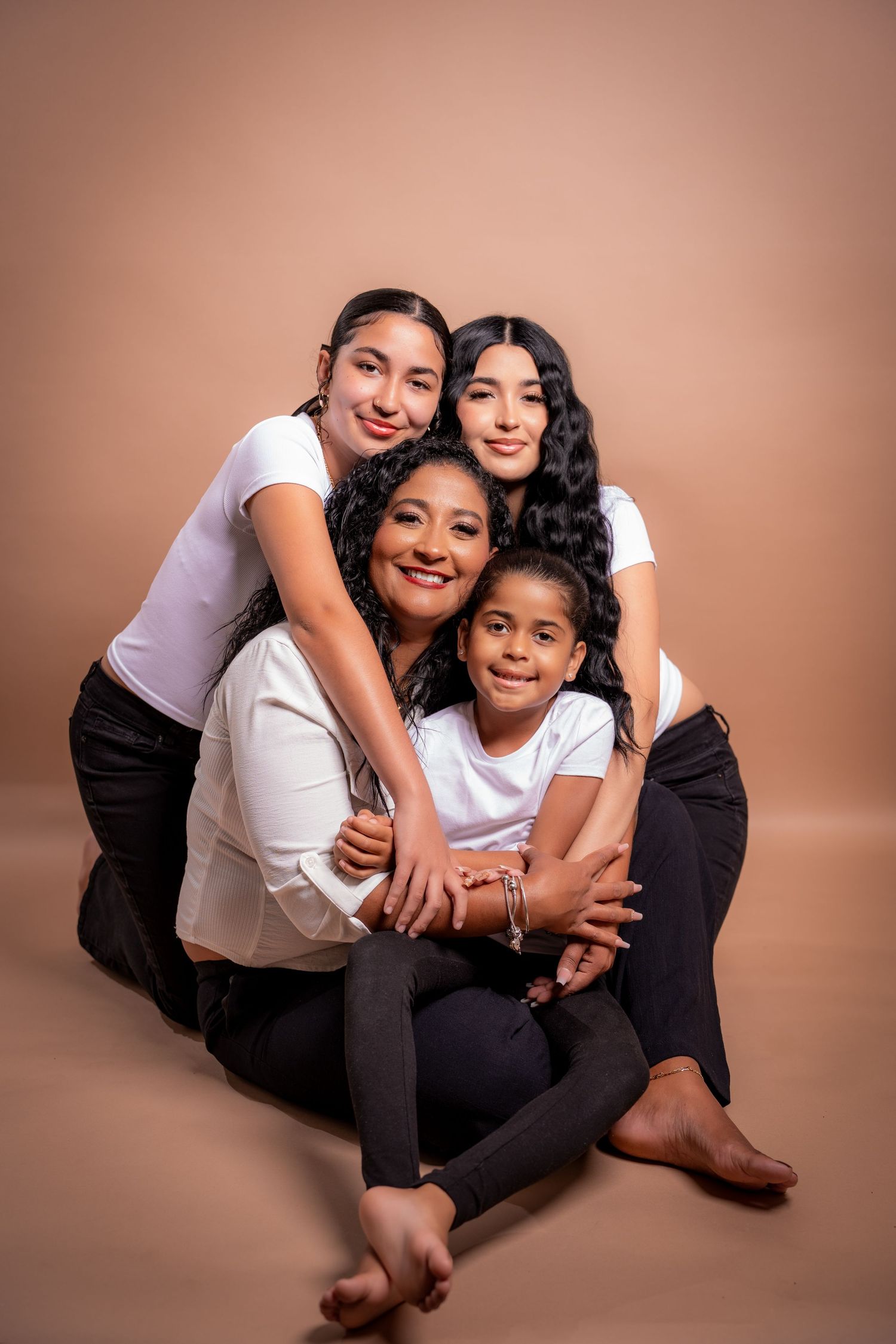 Three people in white and black outfits pose together playfully on the ground in a studio setting with brown backdrop.