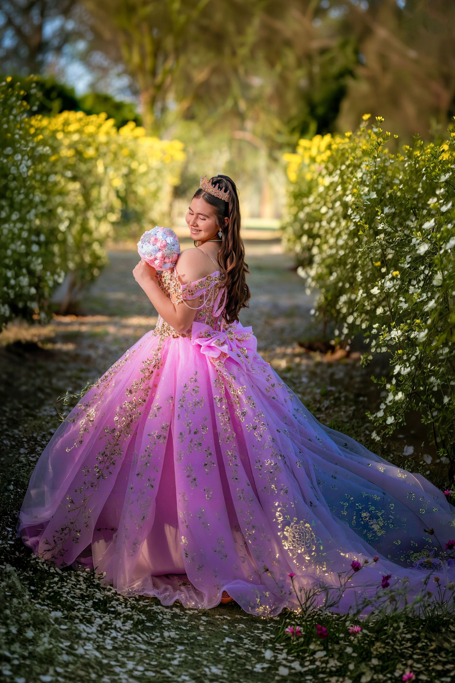 Elegant teen in pink floral quinceañera gown with gold details and tiara walks through a lush garden pathway.
